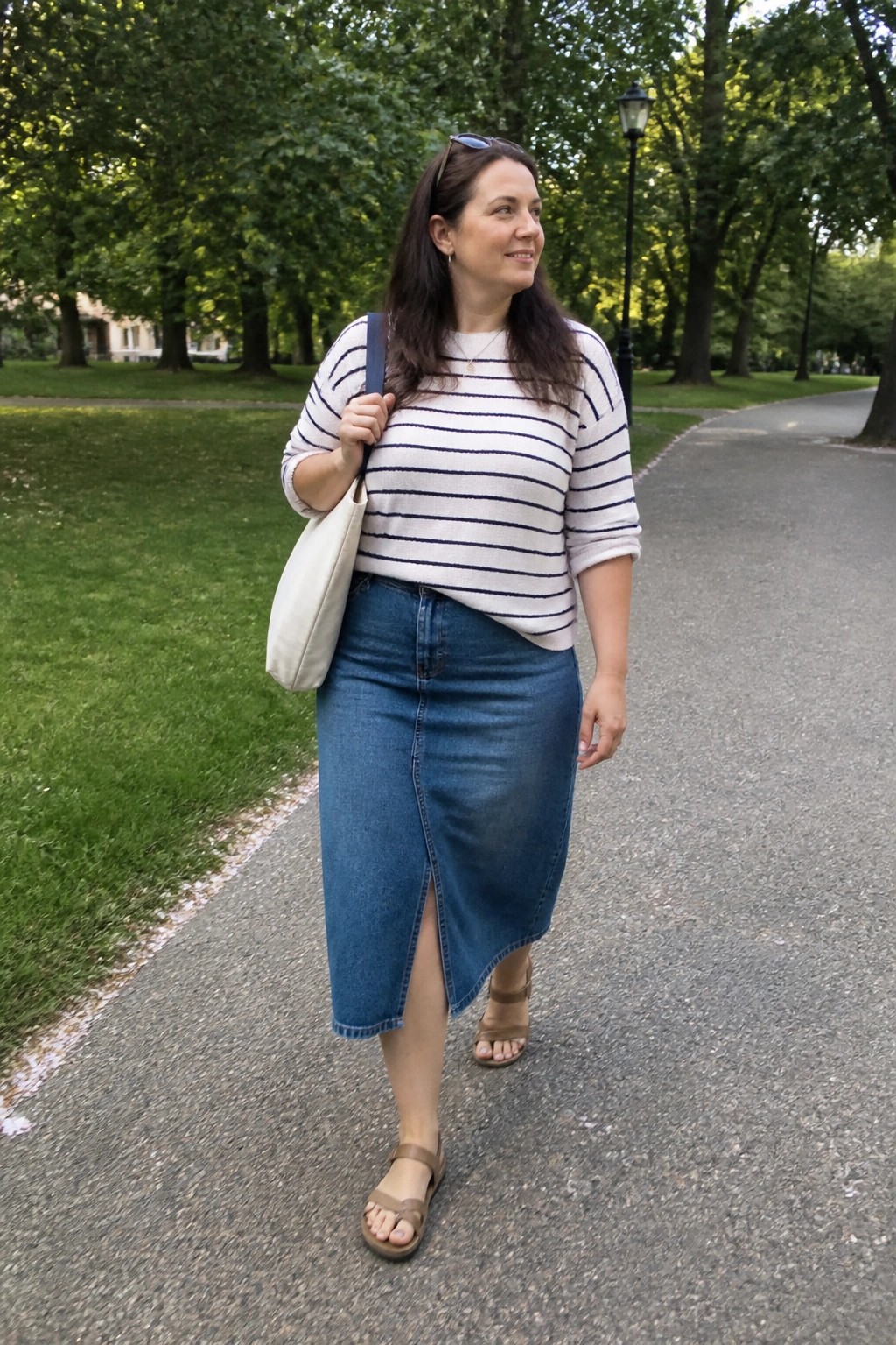 Plus size woman wearing a white and navy striped knit sweater with a blue denim midi skirt featuring a front slit, beige canvas tote, brown strappy sandals, and sunglasses resting on her head while standing on a park path.