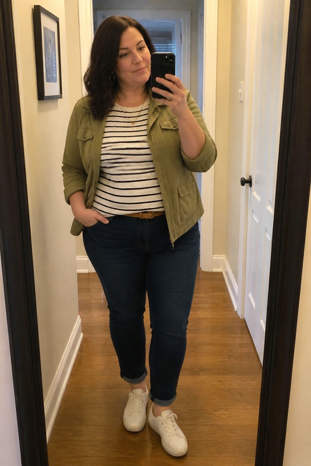 Woman in hallway taking mirror selfie in green jacket over striped shirt and jeans