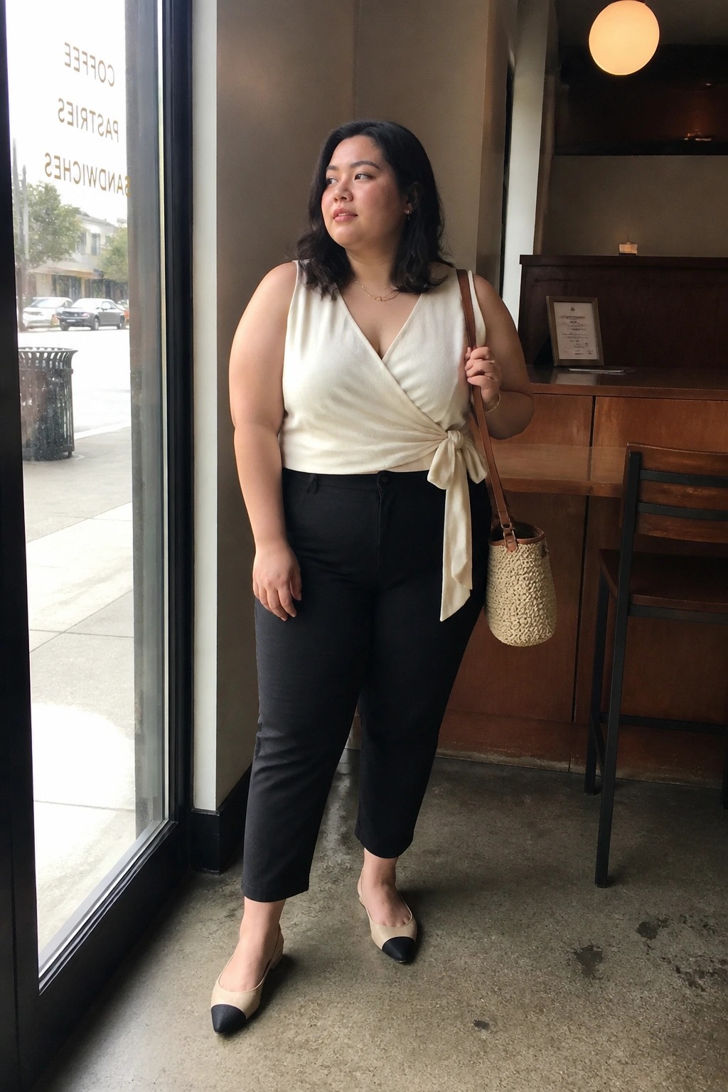 Woman in cream wrap top and black pants with woven bag in cafe