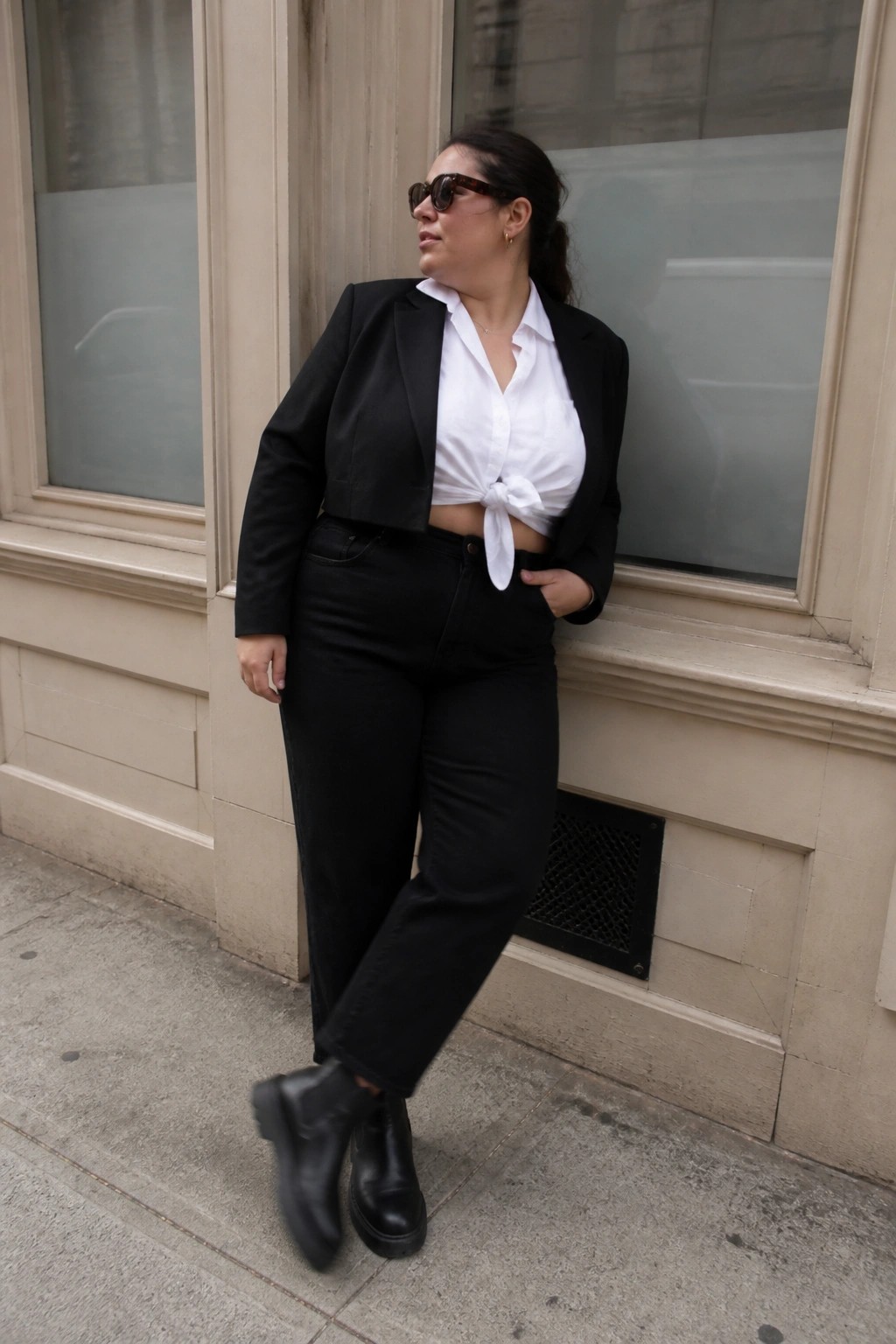 Woman wearing a black cropped blazer over a white button down shirt tied at the waist paired with black jeans and black boots while leaning against a building exterior.