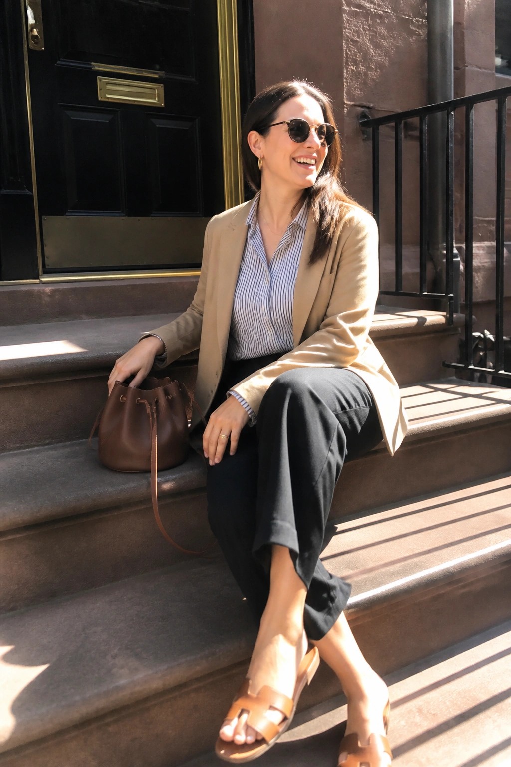 Woman in tan blazer and striped shirt smiling on stone steps with brown bag