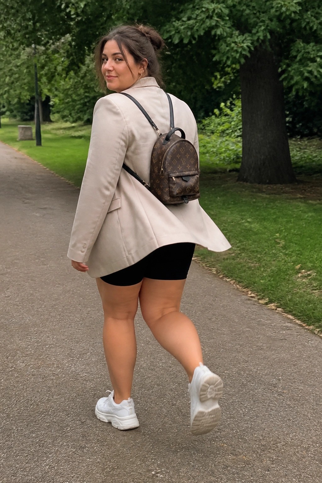 Curvy woman in beige blazer with LV backpack walks on park path