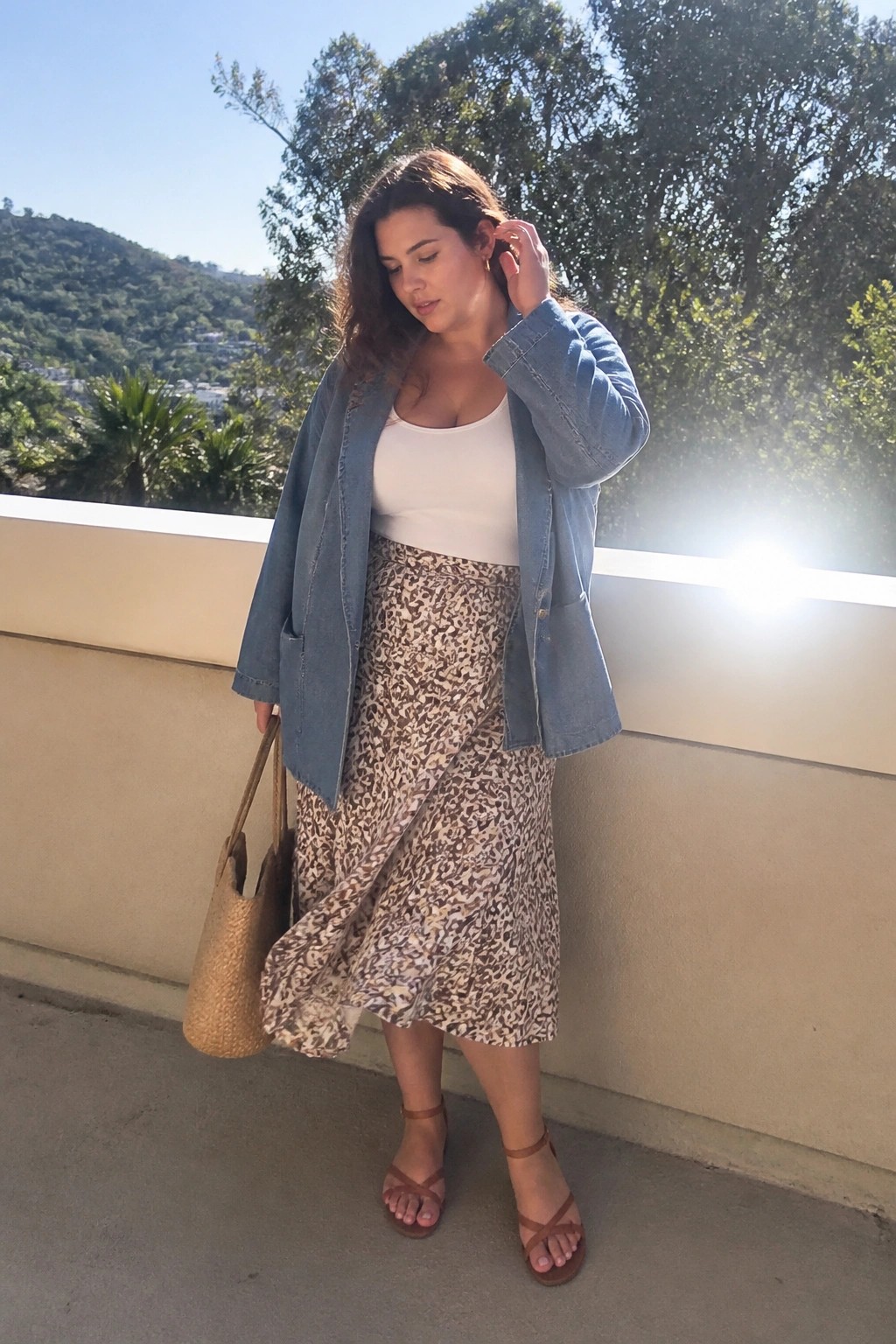 Plus size woman wearing a blue denim jacket over a white tank top with a brown and beige leopard print midi skirt, brown strappy sandals, and carrying a woven straw bag.