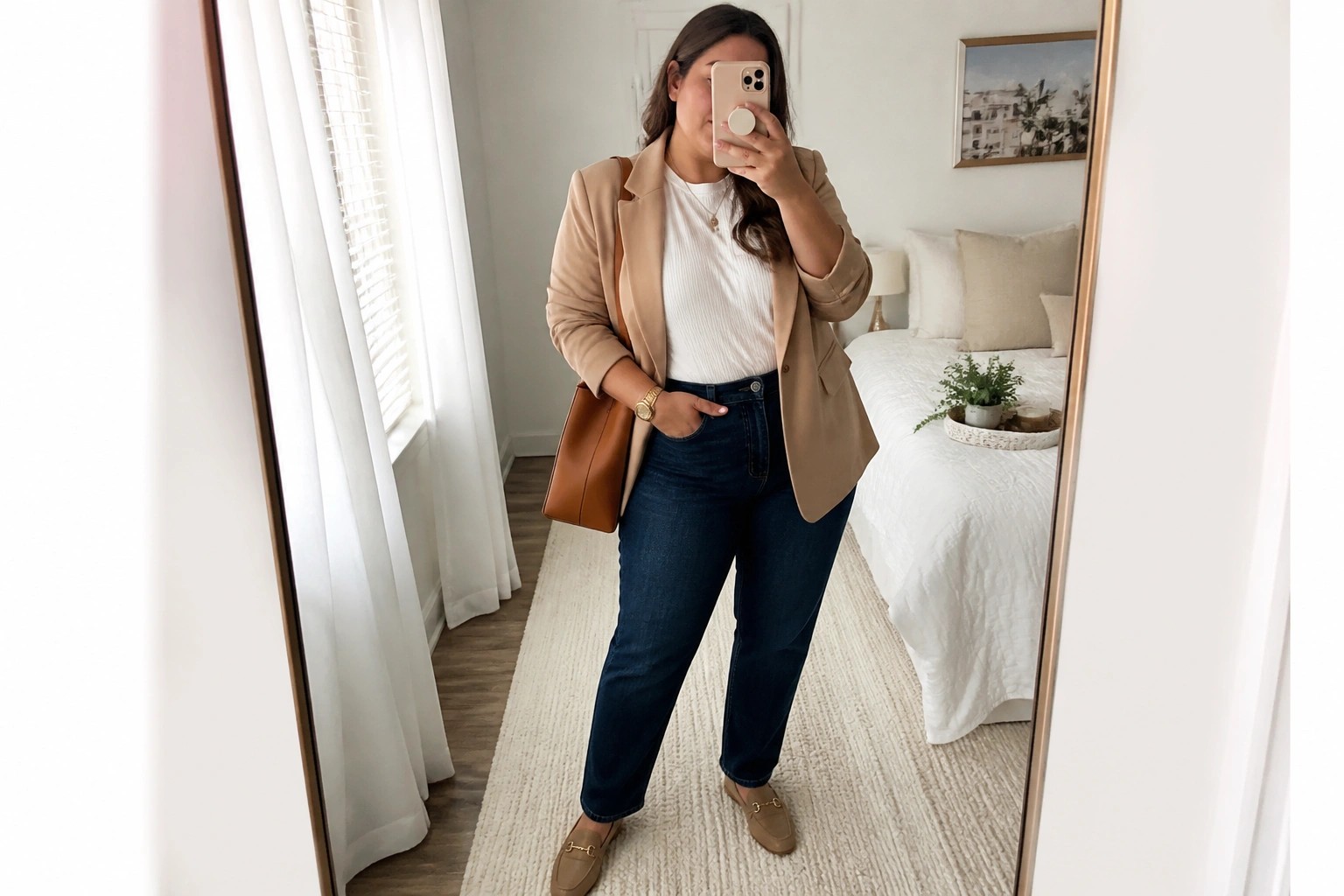 Woman in tan blazer and jeans taking mirror selfie in bedroom