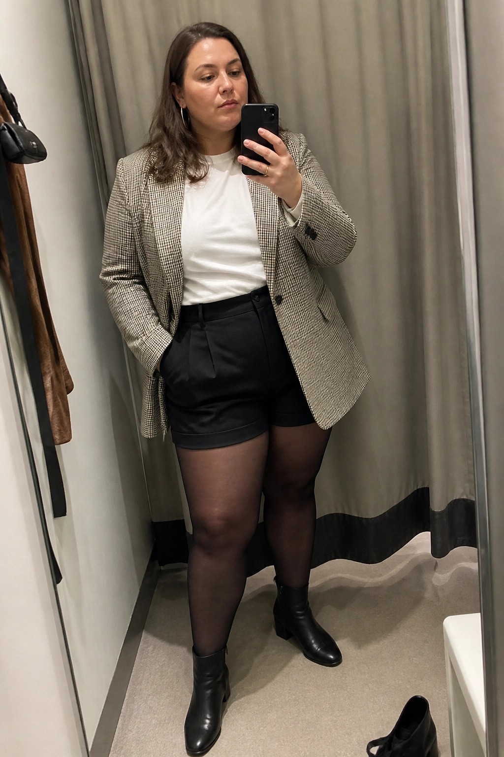 Woman in houndstooth blazer and black shorts takes mirror selfie in fitting room
