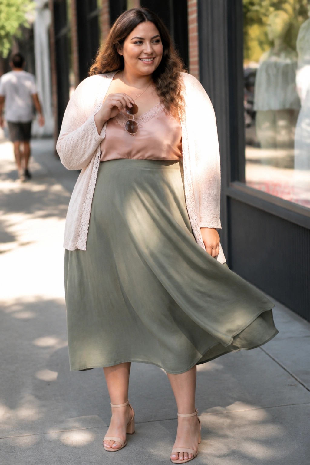 Smiling woman in sage green skirt, pink camisole, and beige cardigan on sidewalk.