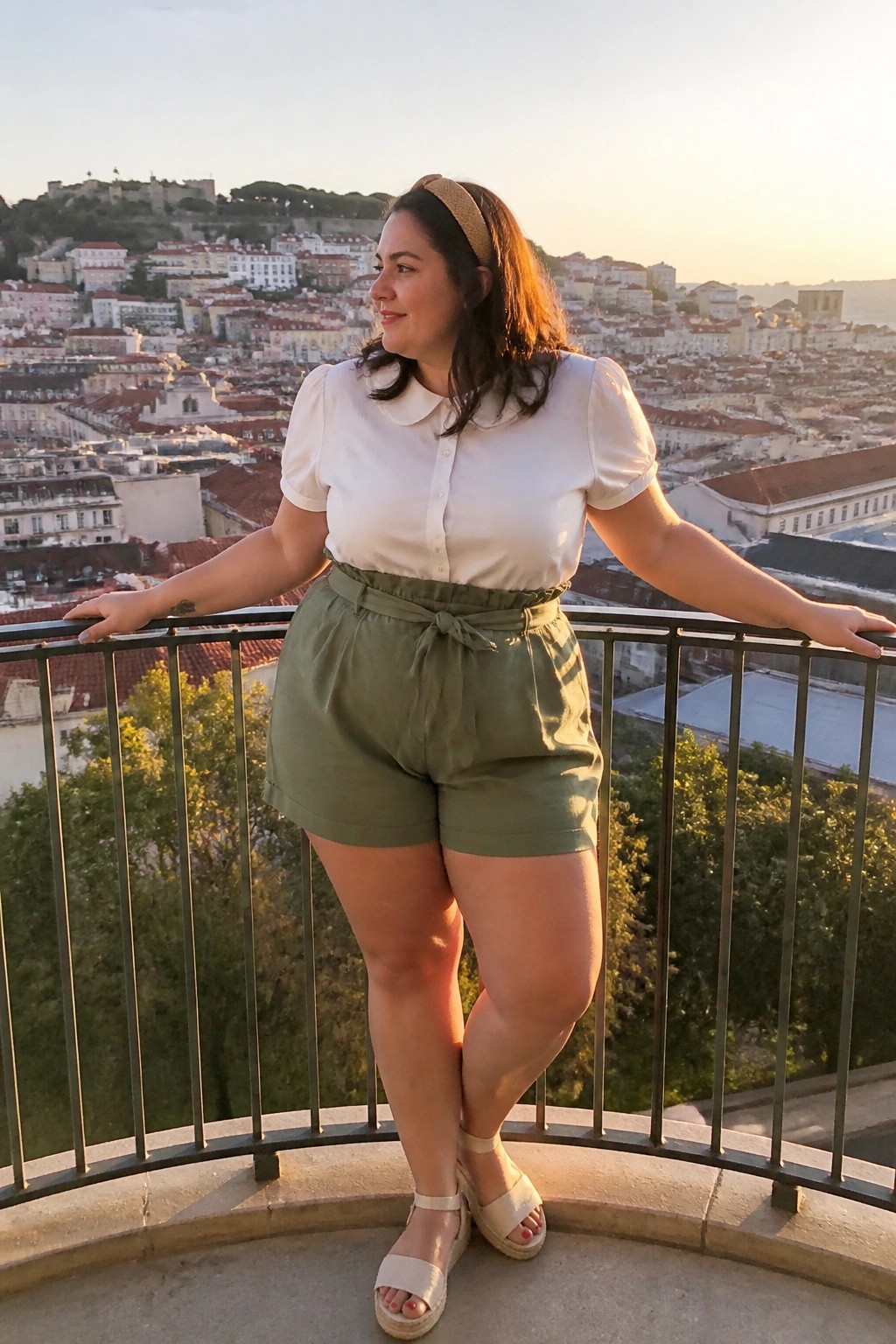 Woman in white blouse and green shorts on balcony overlooking city at sunset