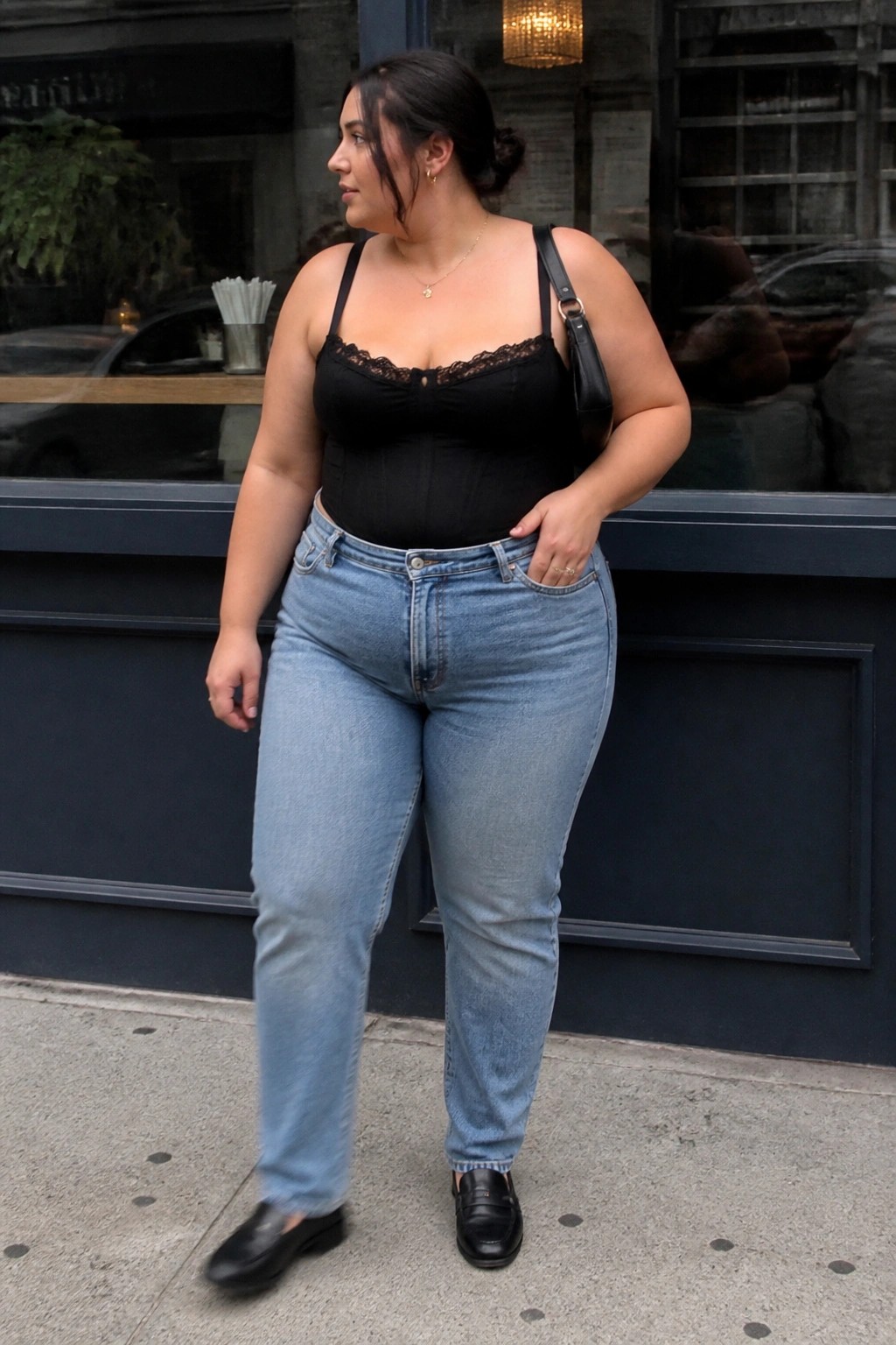Plus size woman wearing a black lace trimmed corset top tucked into light blue high waisted jeans with black loafers and a shoulder bag.