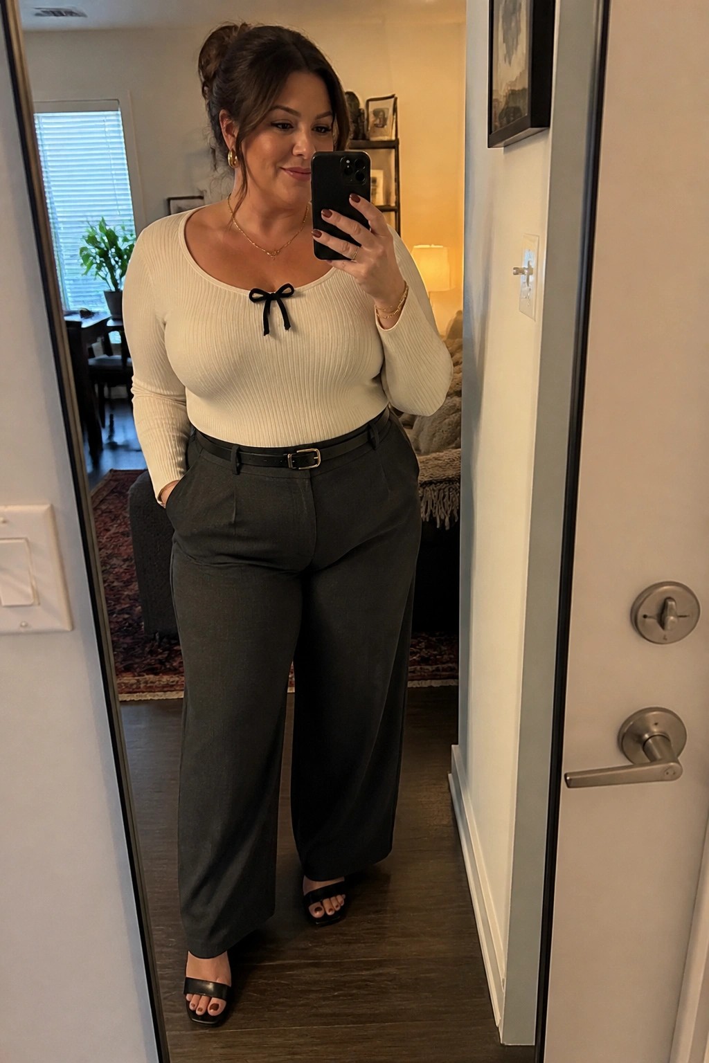 Plus size woman wearing a cream ribbed long sleeve top with black bow detail paired with dark wide leg trousers and a black belt in a mirror selfie.