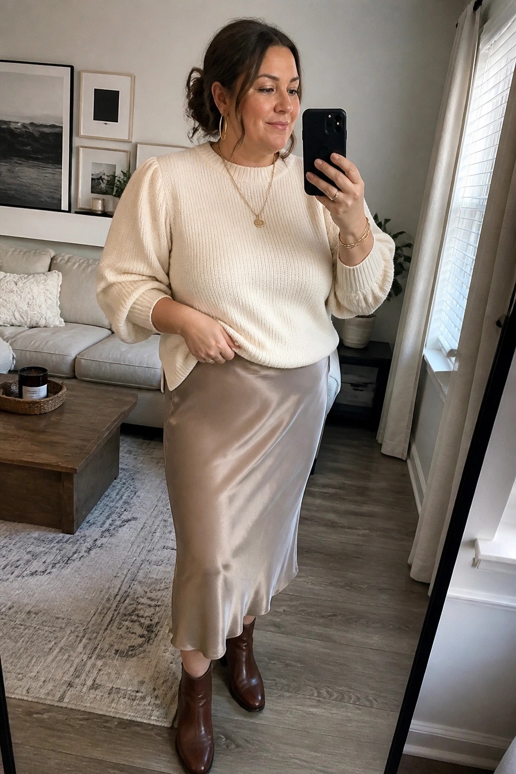 Plus size woman in cream puff sleeve knit sweater tucked slightly into beige satin midi skirt with brown leather ankle boots and layered gold necklaces and bracelets taking a mirror selfie.