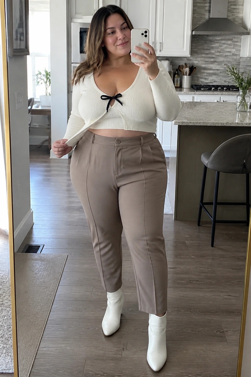 Woman in white cropped cardigan with black bow, beige pants, and white boots taking mirror selfie