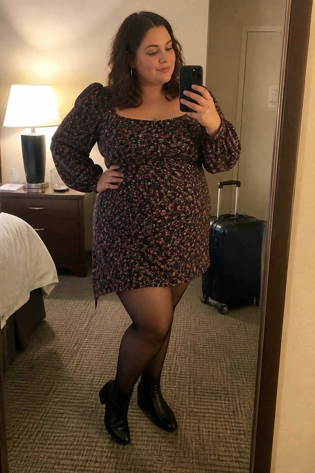 Curvy woman in floral mini dress taking mirror selfie in hotel room with suitcase
