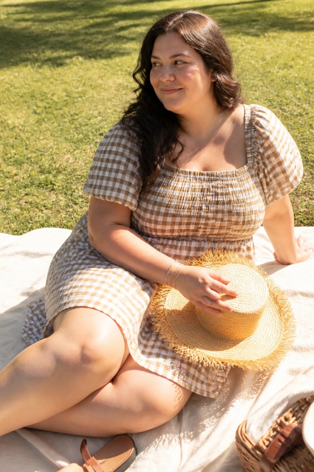 Plus size woman wearing a brown gingham check mini dress with smocked square neckline and puff sleeves while seated on a blanket holding a straw hat.