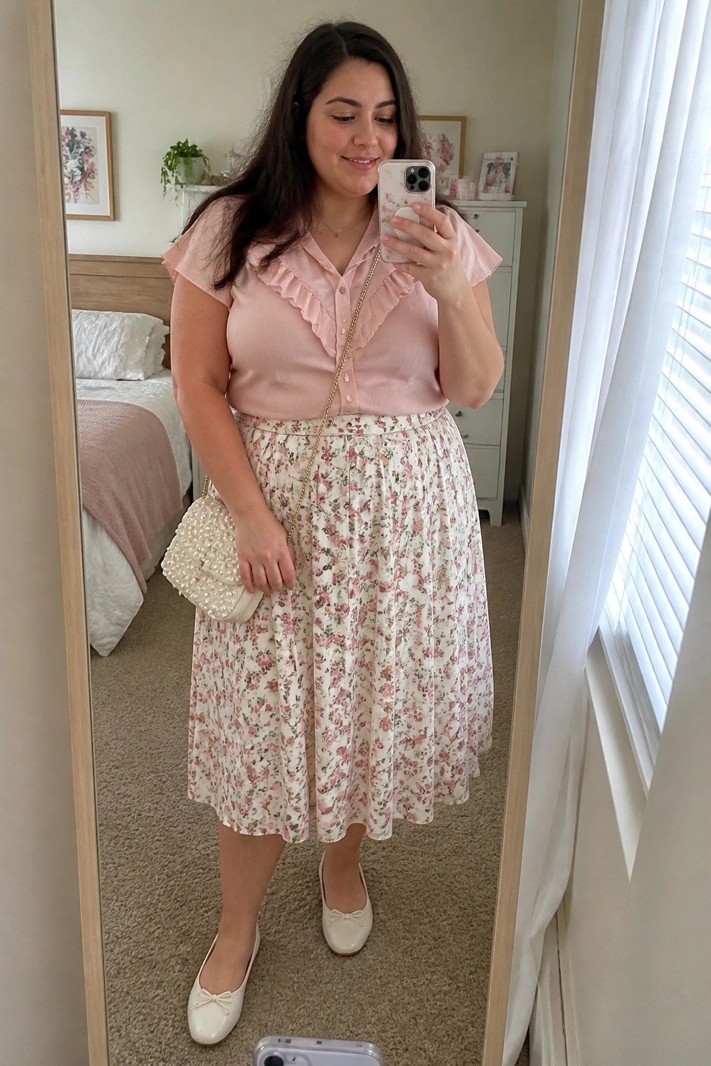 Plus size woman wearing a pink ruffled blouse tucked into a cream floral midi skirt with a pearl chain bag and white bow flats while taking a mirror selfie.