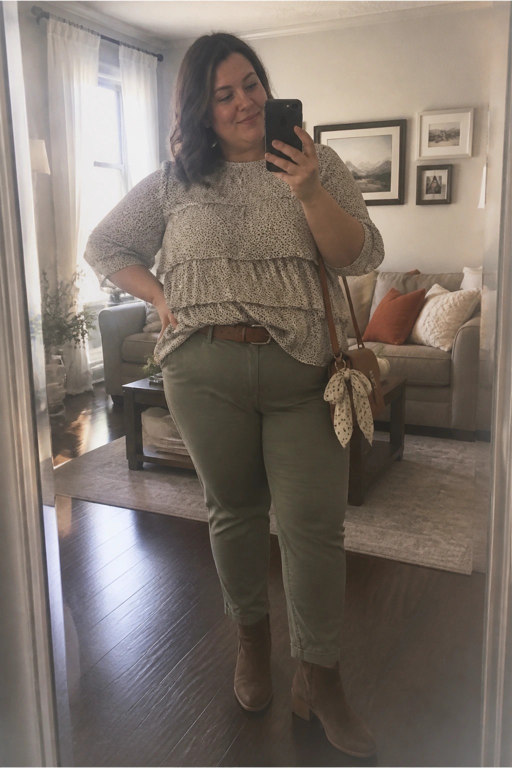 Plus size woman wearing a cream and black spotted tiered ruffle blouse tucked into olive green pants with a brown belt, brown crossbody bag featuring a polka dot bow, and matching brown ankle boots in a living room mirror selfie.