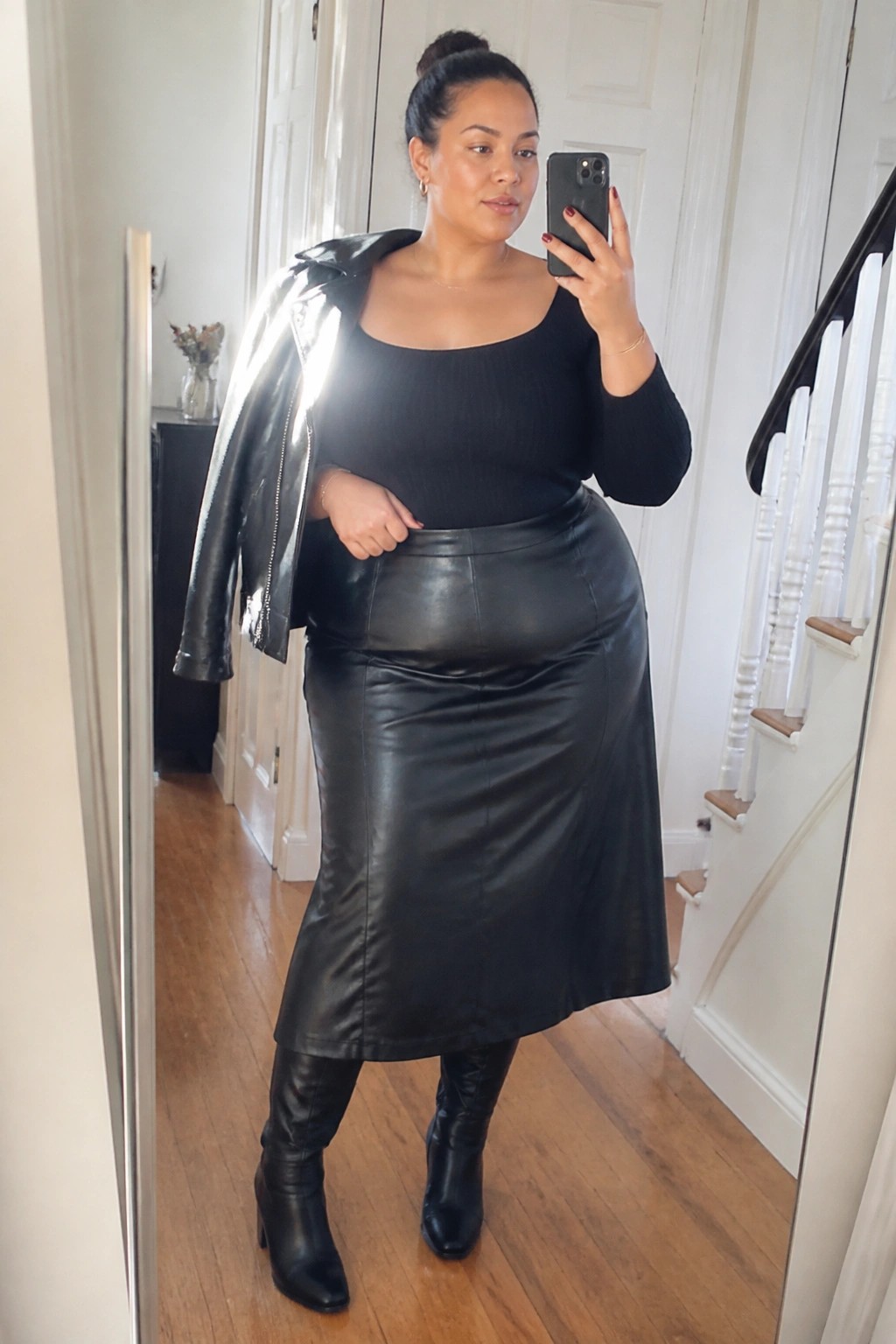 Woman in black top and leather skirt taking mirror selfie with jacket over shoulder