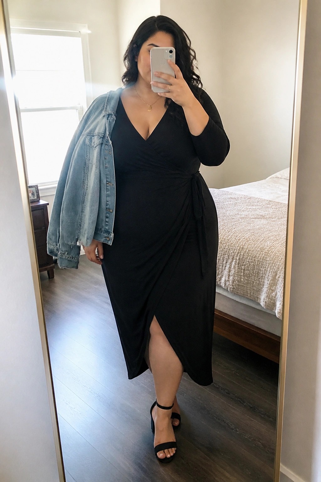 Woman in black wrap dress with denim jacket taking mirror selfie in bedroom