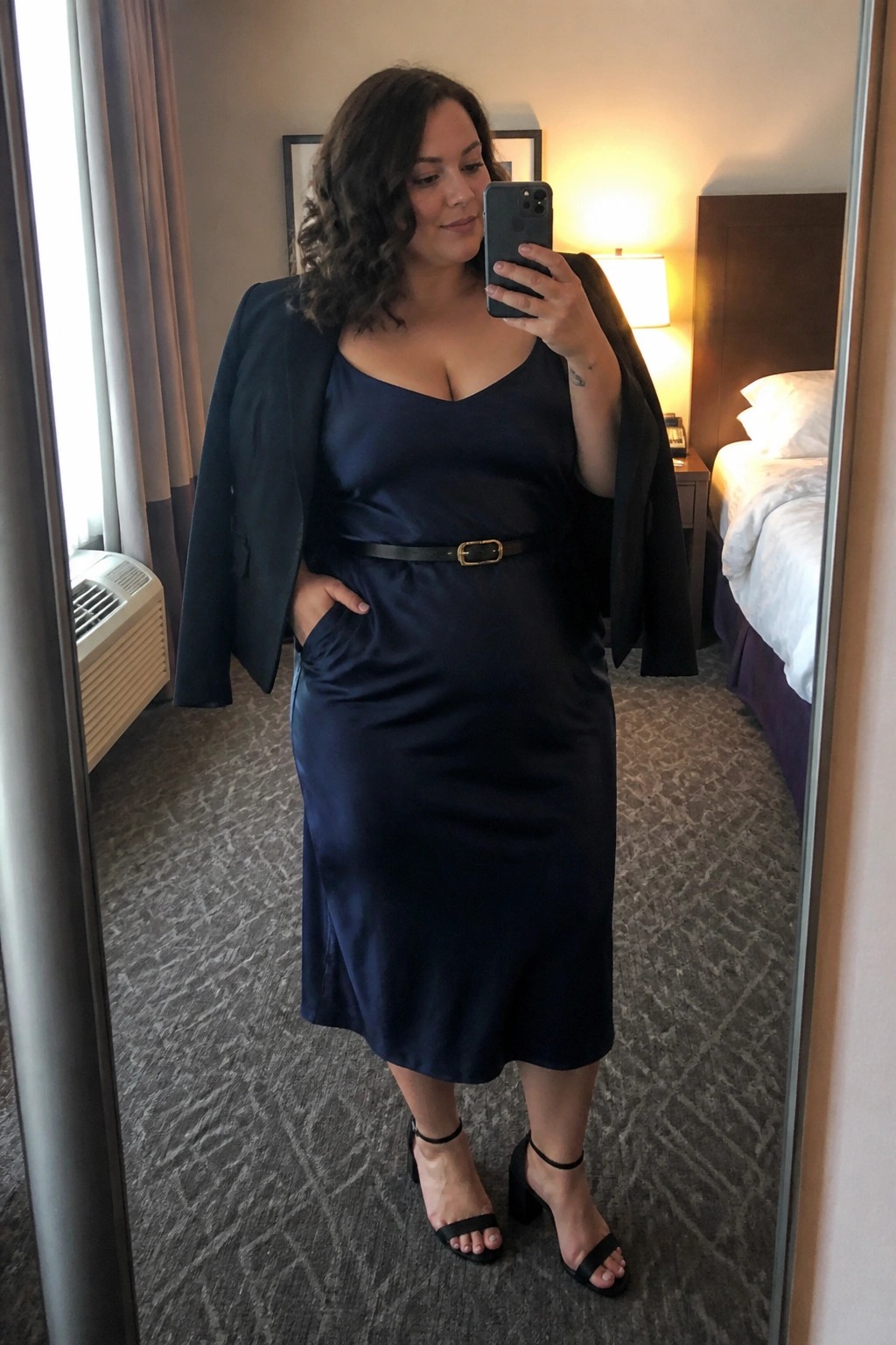 Woman in navy satin dress taking mirror selfie in hotel room