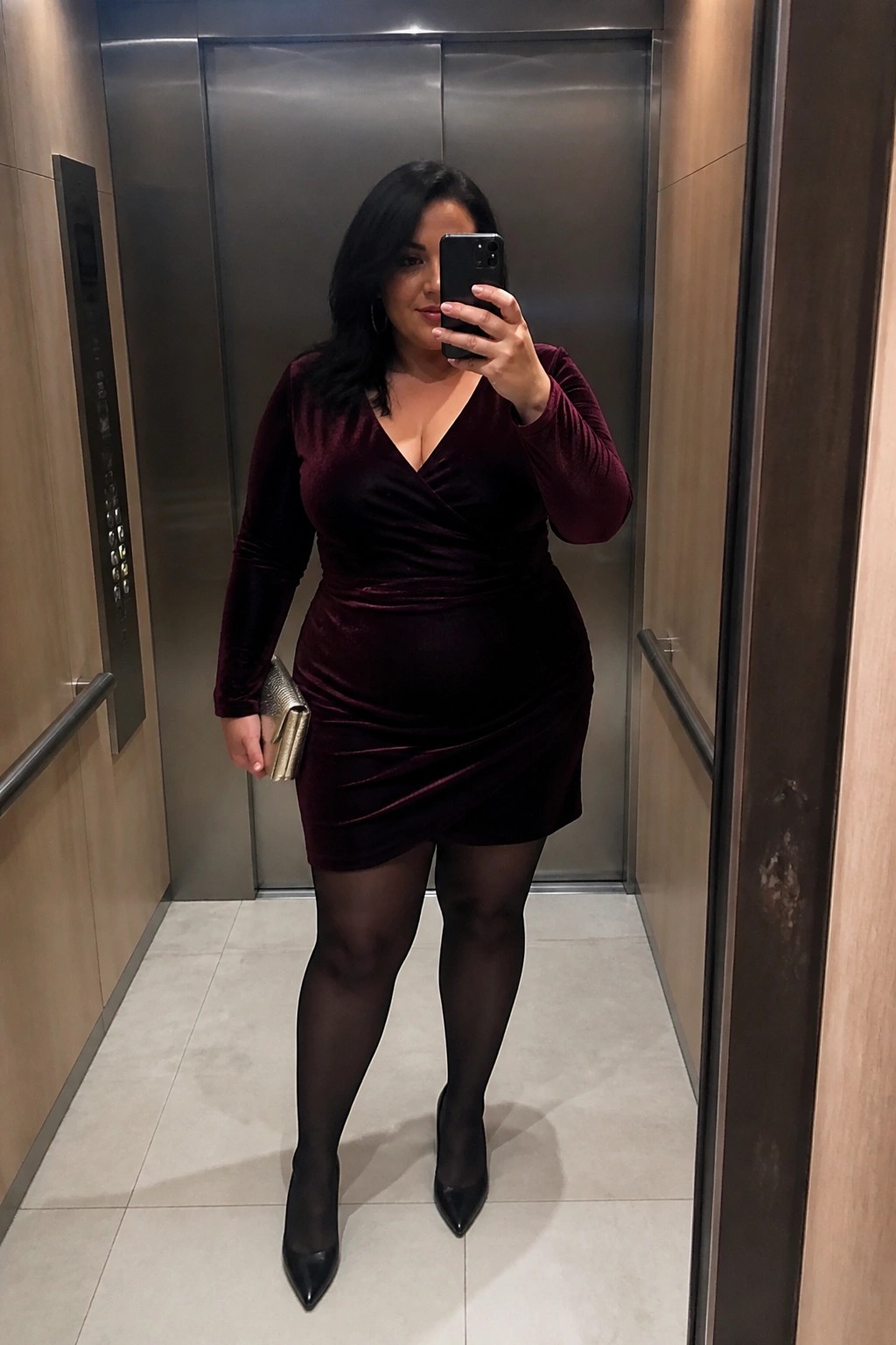 A plus size woman wearing a burgundy velvet wrap mini dress with black tights and heels holding a gold clutch in an elevator mirror selfie.