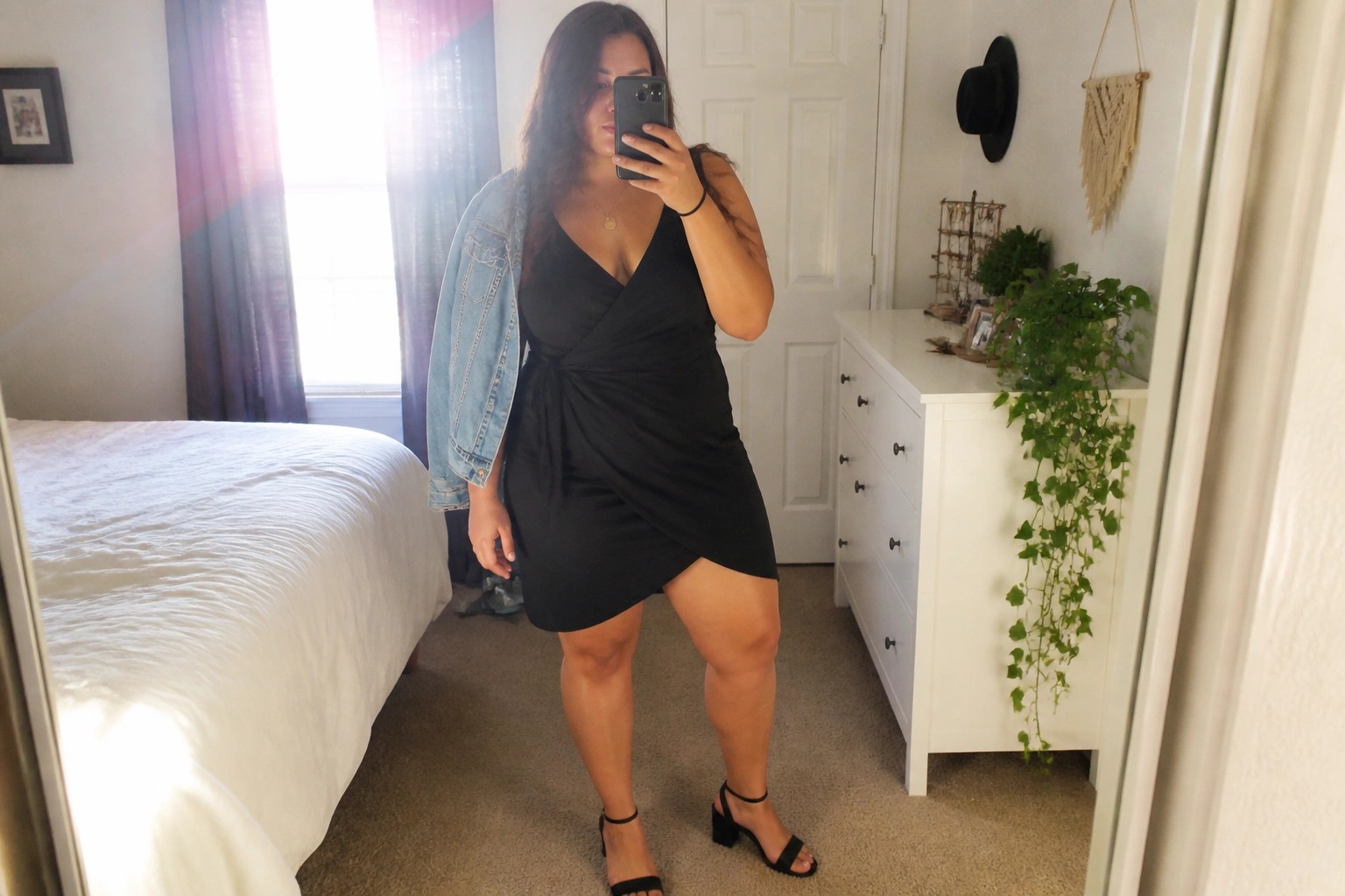 Woman in black wrap dress with denim jacket taking mirror selfie in bedroom