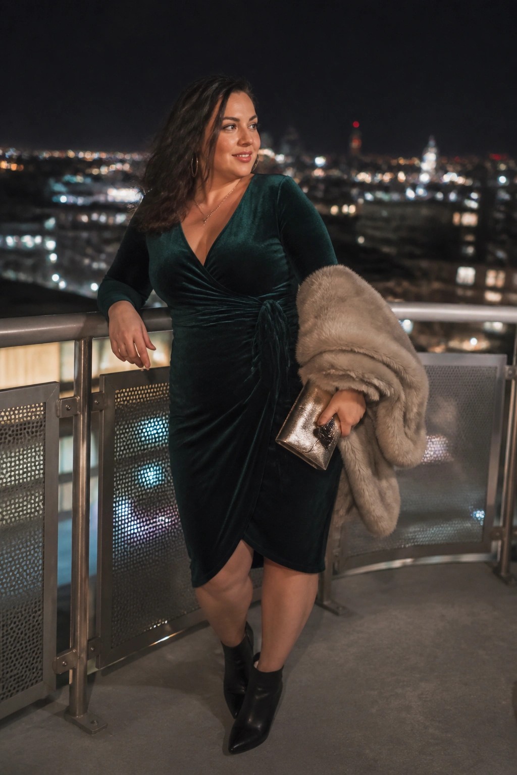 Woman in green velvet dress holding fur coat and clutch on nighttime city balcony