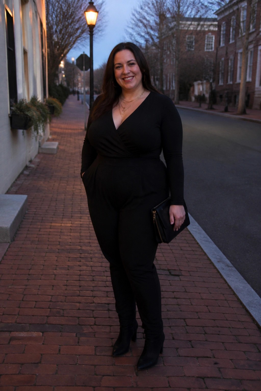 Woman in black jumpsuit holding clutch on brick sidewalk at dusk