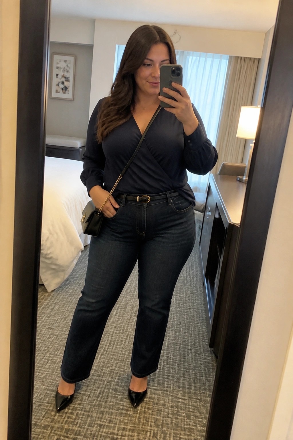 Woman in navy wrap top and jeans taking mirror selfie in hotel room