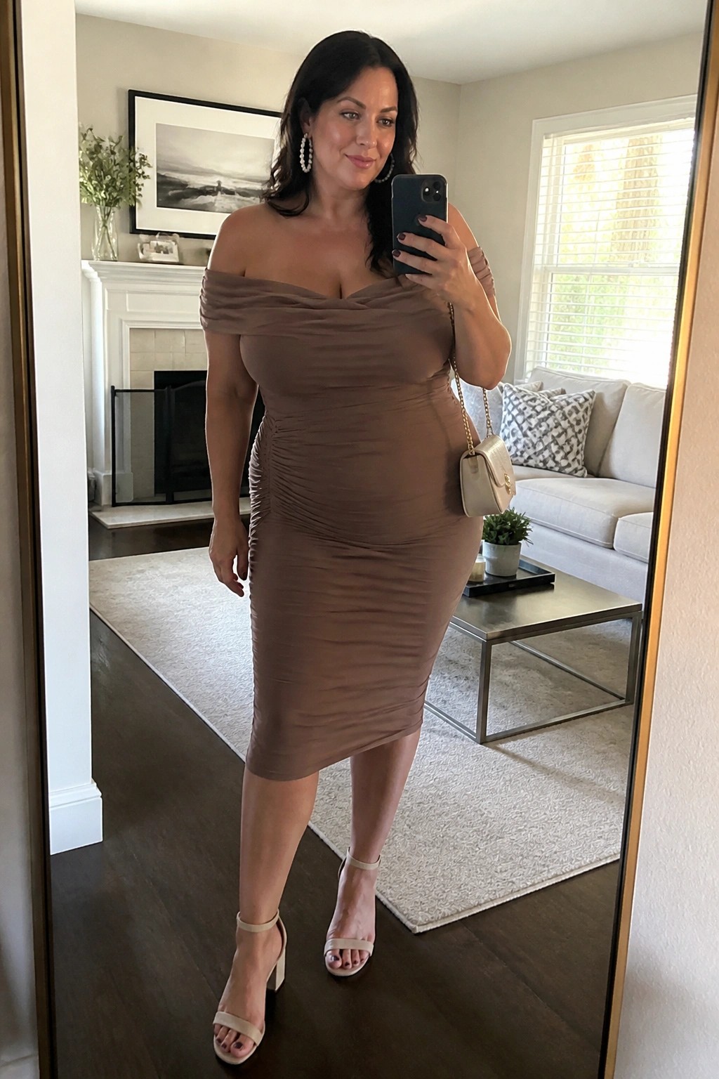 Woman in taupe off-shoulder ruched dress taking mirror selfie in living room