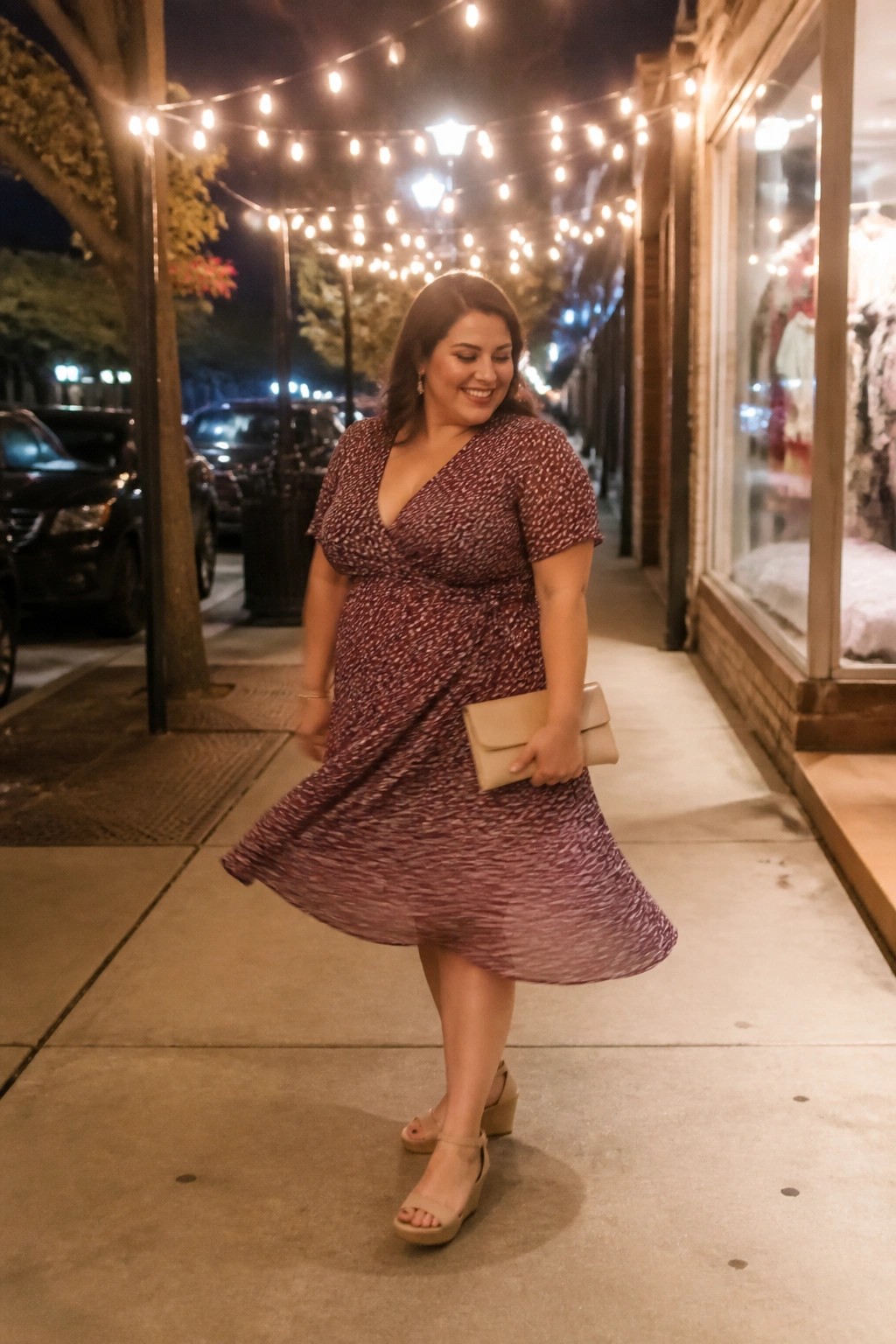 Plus size woman in a burgundy patterned wrap dress paired with a beige clutch and matching wedge sandals.