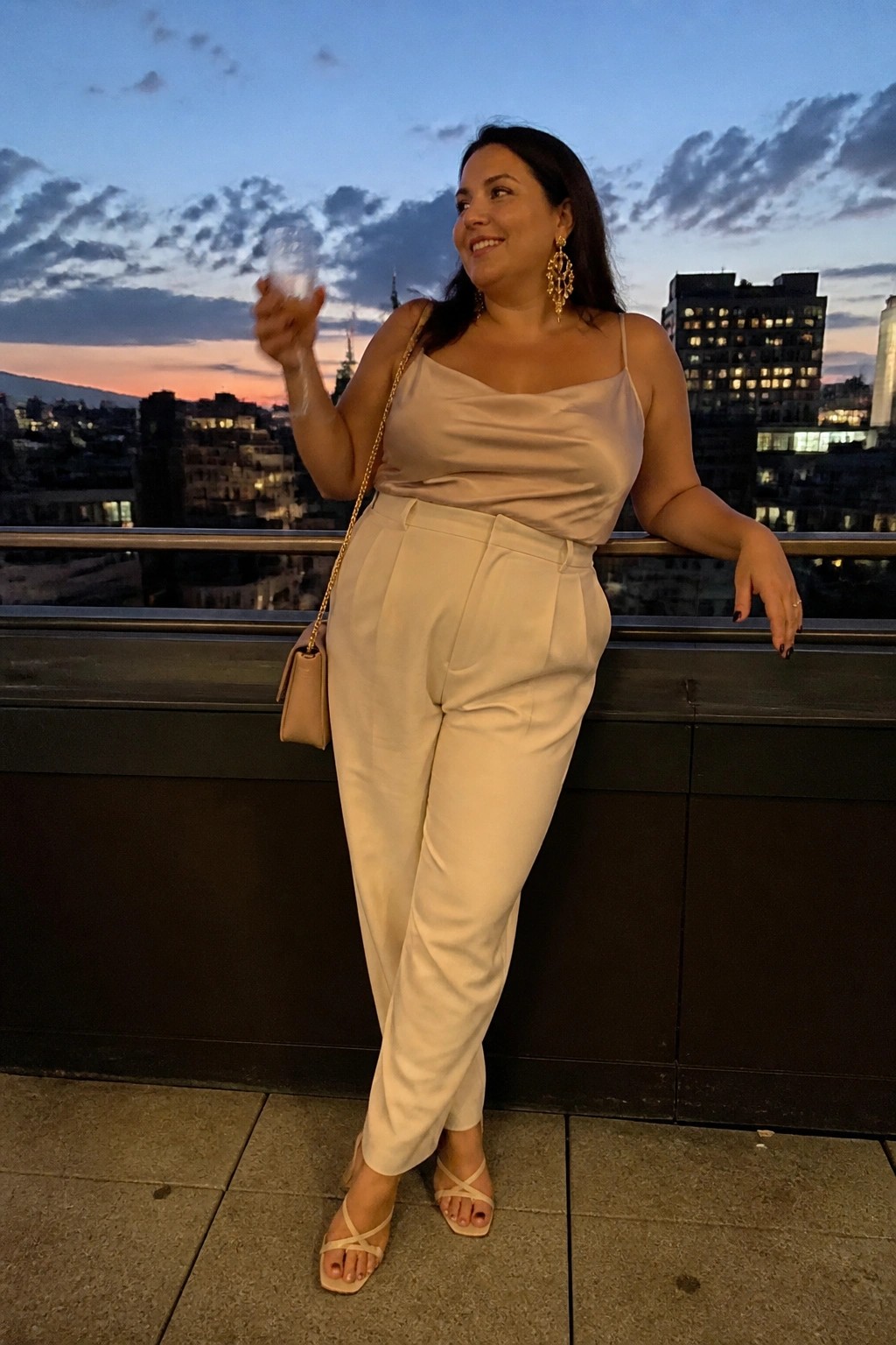 Woman in beige satin top and cream pants holding wine glass on city balcony at dusk