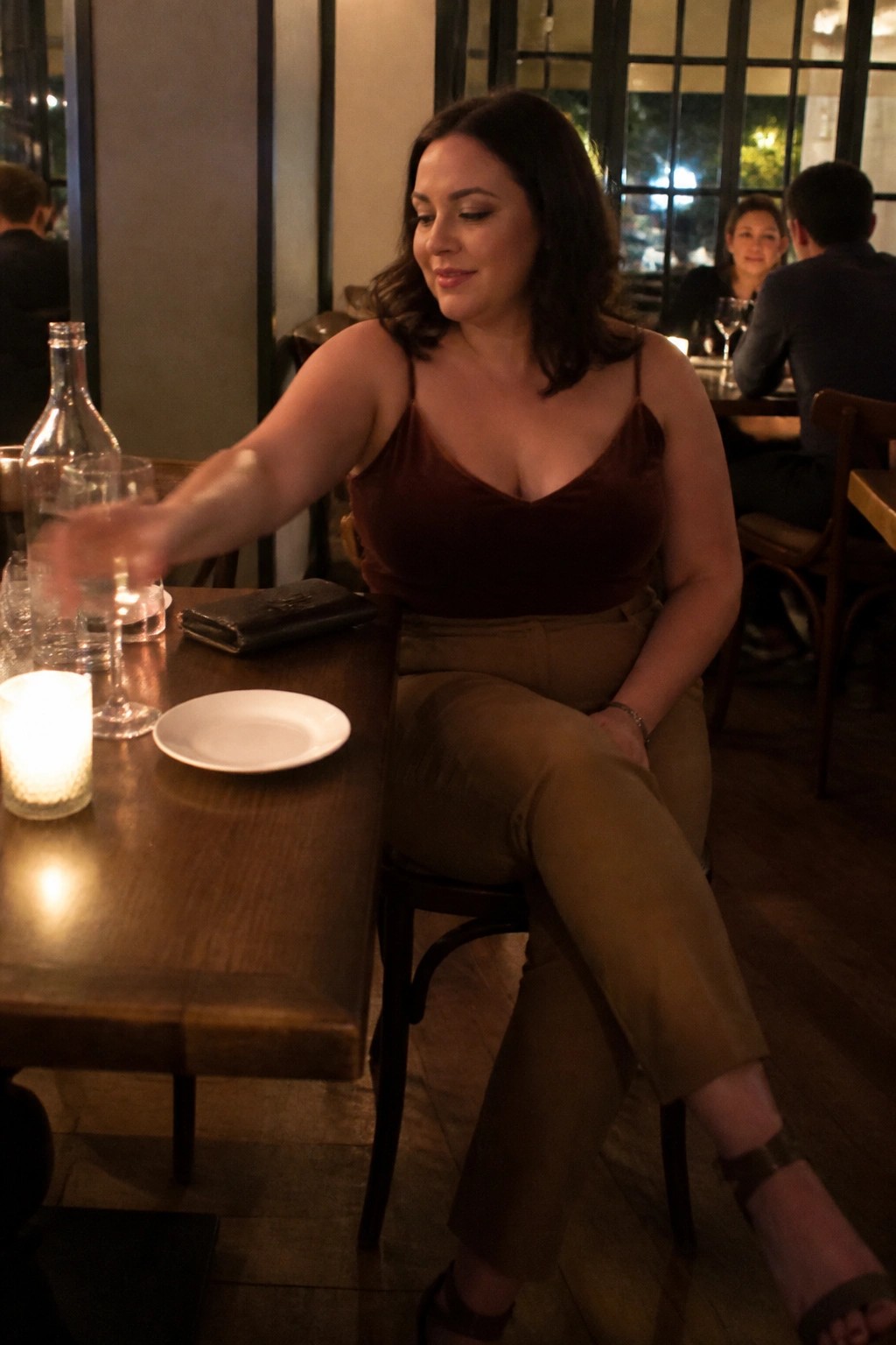 Plus size woman in brown velvet camisole and matching brown trousers seated at a wooden restaurant table with a lit candle and wine glasses nearby.