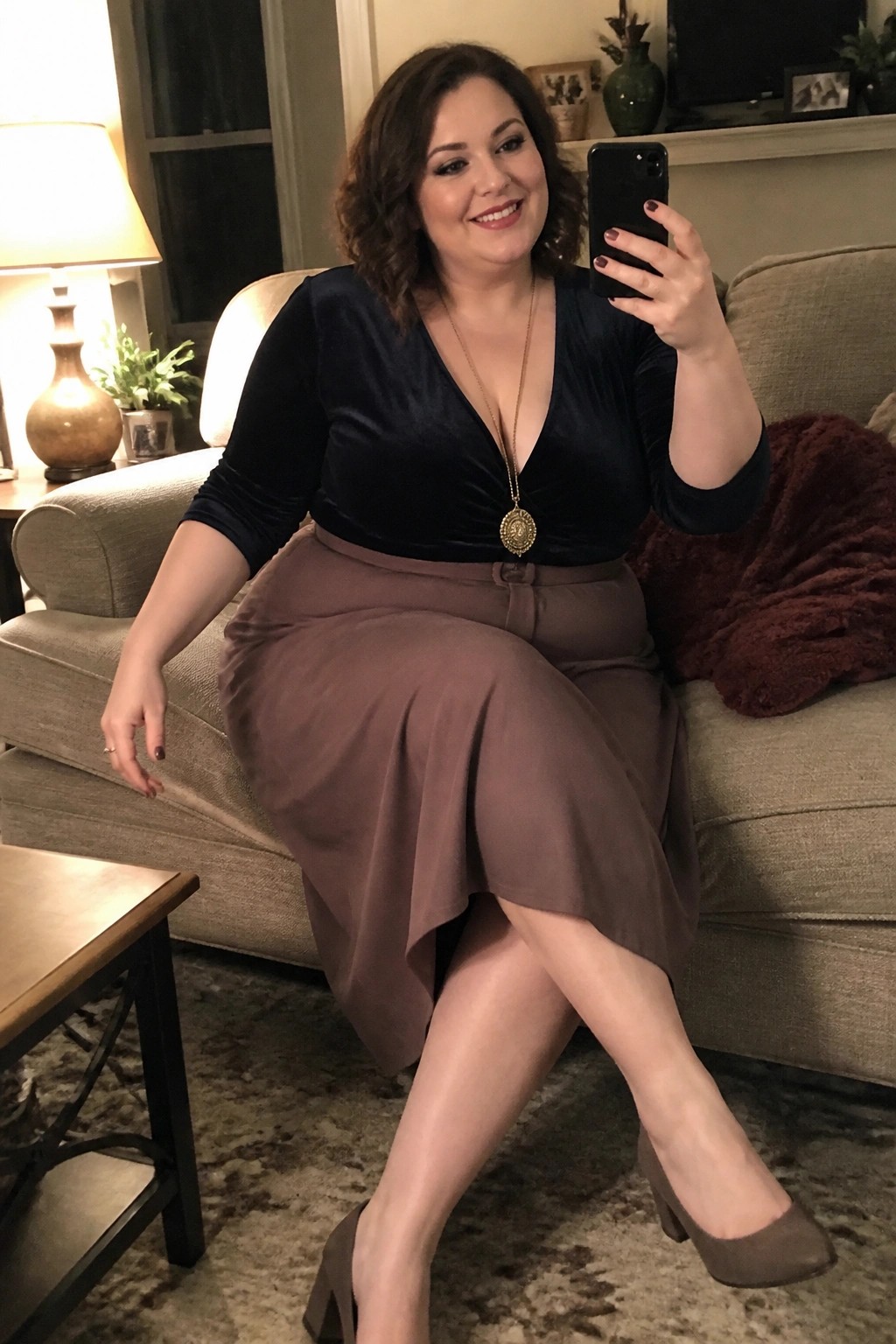 Woman in navy velvet top and brown skirt taking selfie on couch