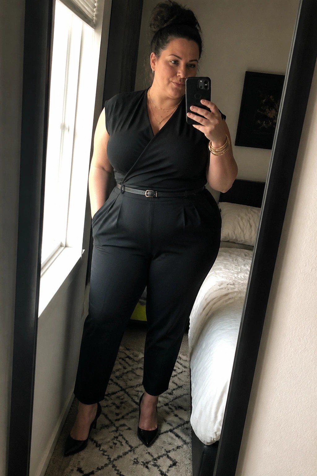 Woman in black wrap top and trousers taking mirror selfie in bedroom