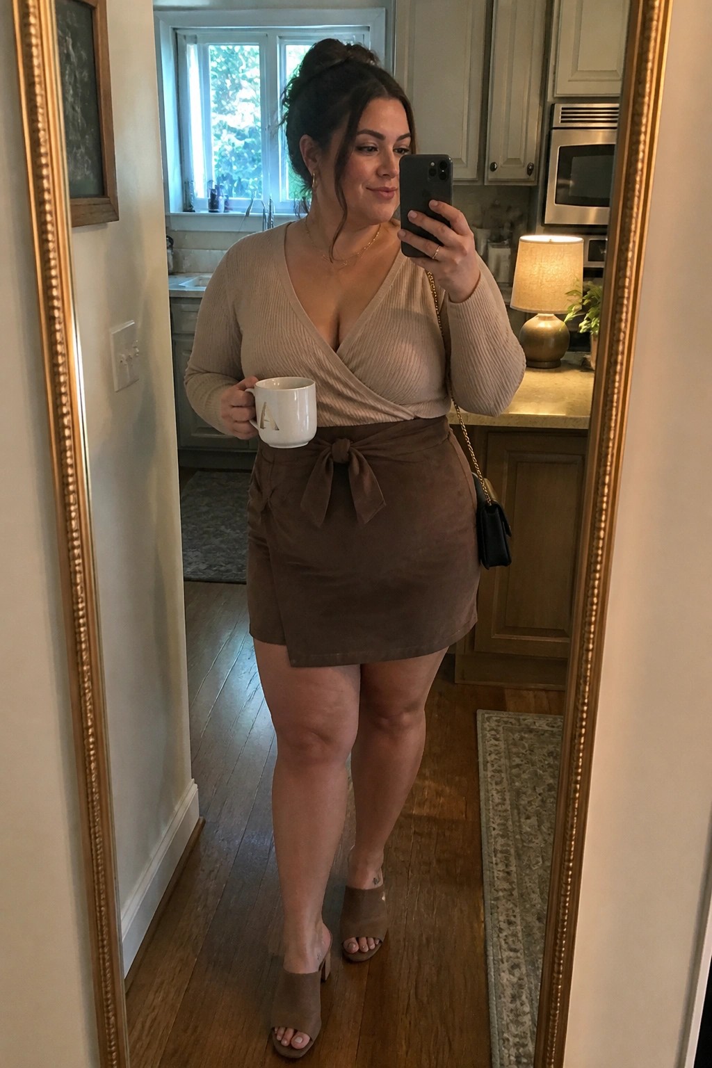 Woman in beige wrap top and brown suede skirt taking kitchen mirror selfie with mug