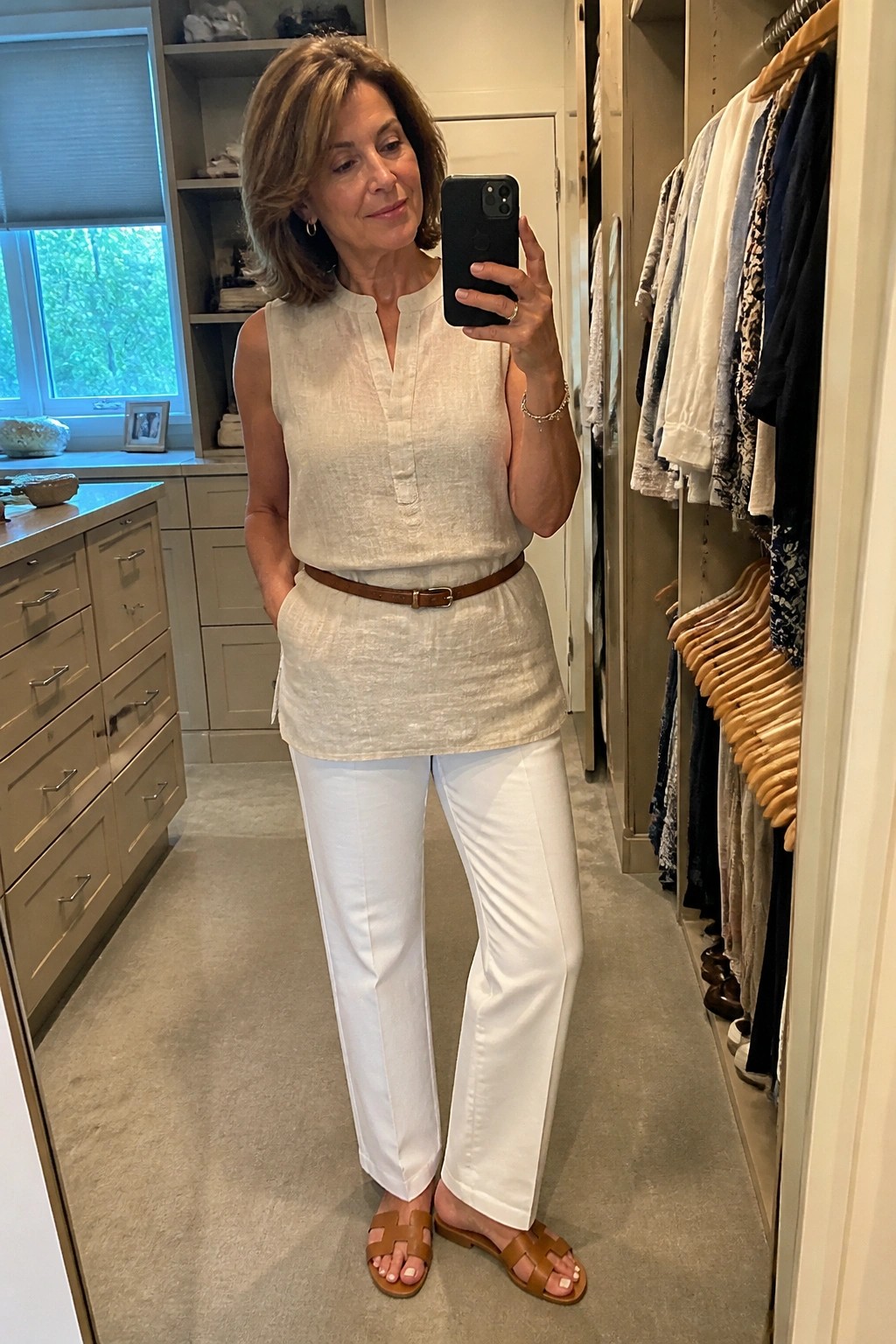 Woman in beige linen top and white pants taking mirror selfie in closet