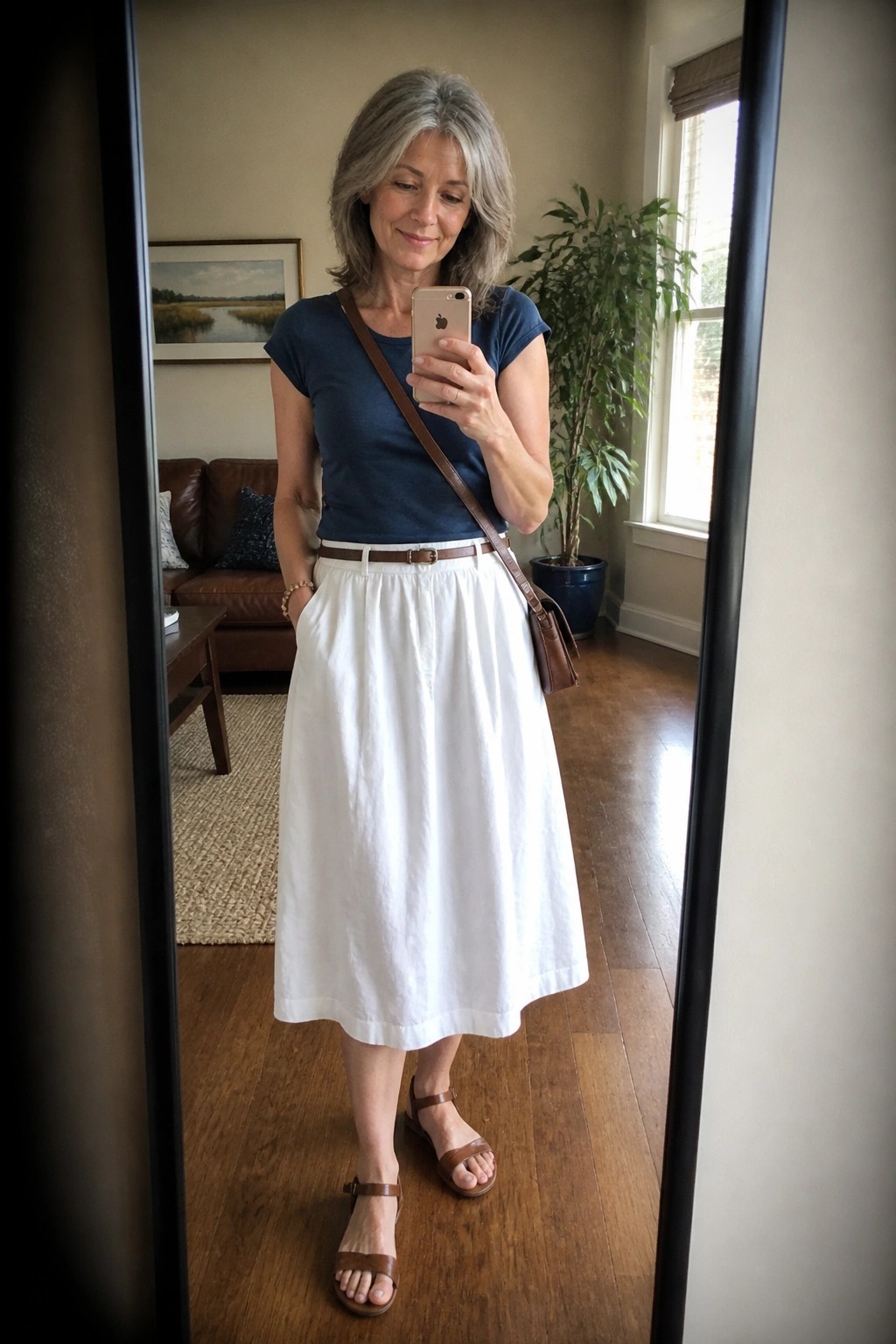 Woman wearing navy blue short sleeve top tucked into white midi skirt with brown belt, brown leather sandals and crossbody bag while taking a mirror selfie.