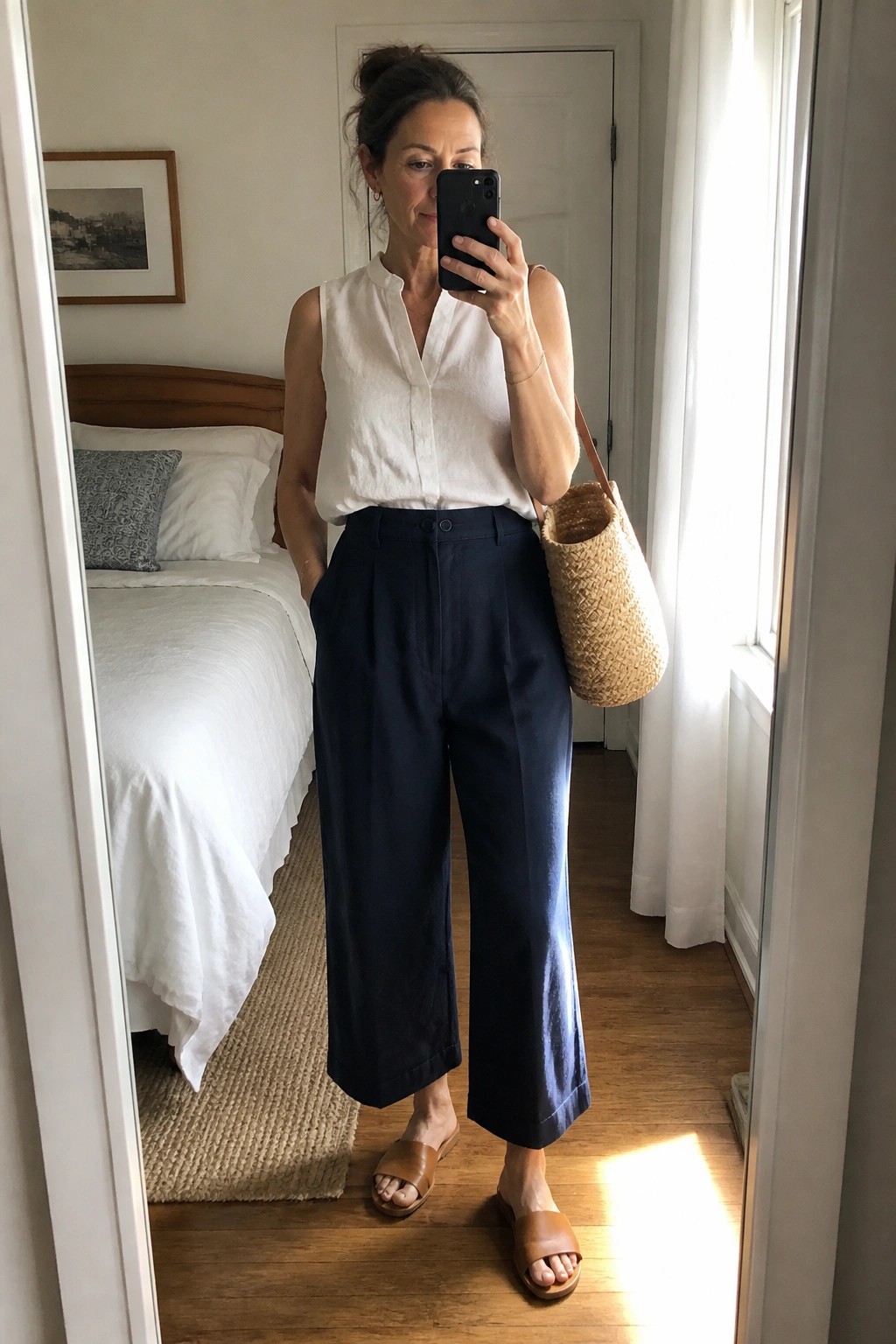 Woman in white sleeveless top and navy wide-leg pants taking bedroom mirror selfie with straw bag