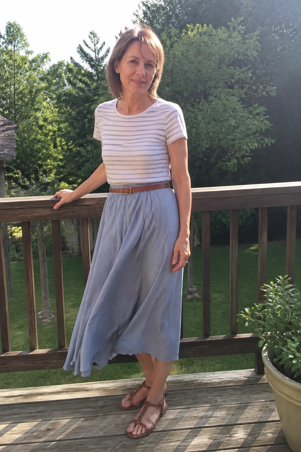 Woman in striped tee and blue skirt leaning on wooden deck railing amid trees