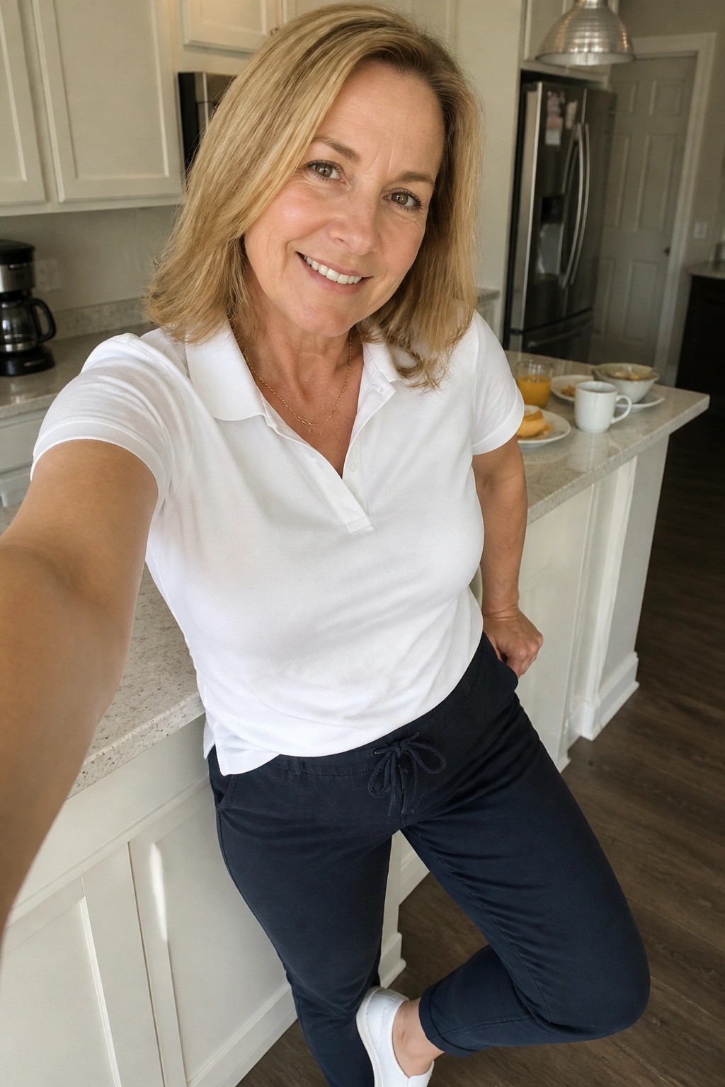 Smiling blonde woman in white polo shirt and navy pants taking kitchen selfie