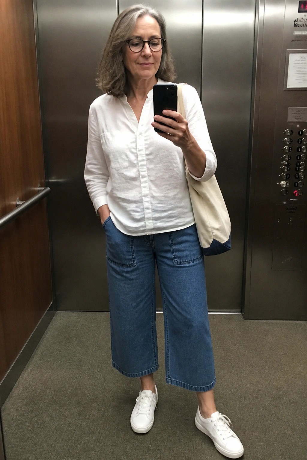 Woman over 50 wearing a white linen button down shirt tucked into blue wide leg cropped denim pants with white sneakers and a beige tote bag while holding a phone in an elevator.