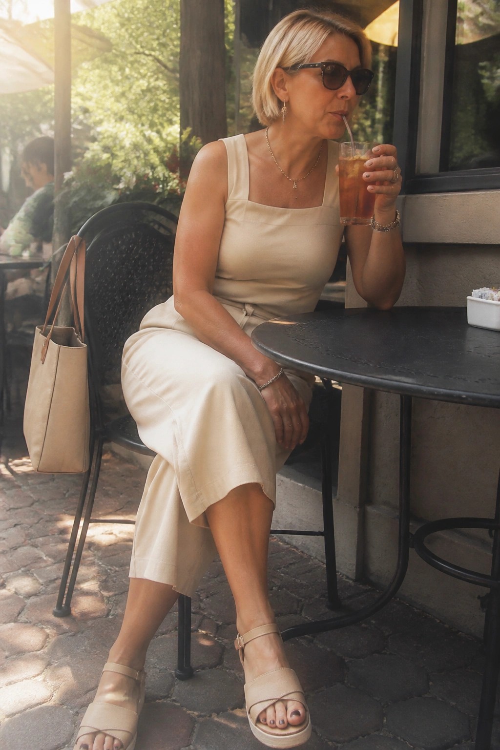 Blonde woman in beige outfit sipping iced drink at outdoor cafe table