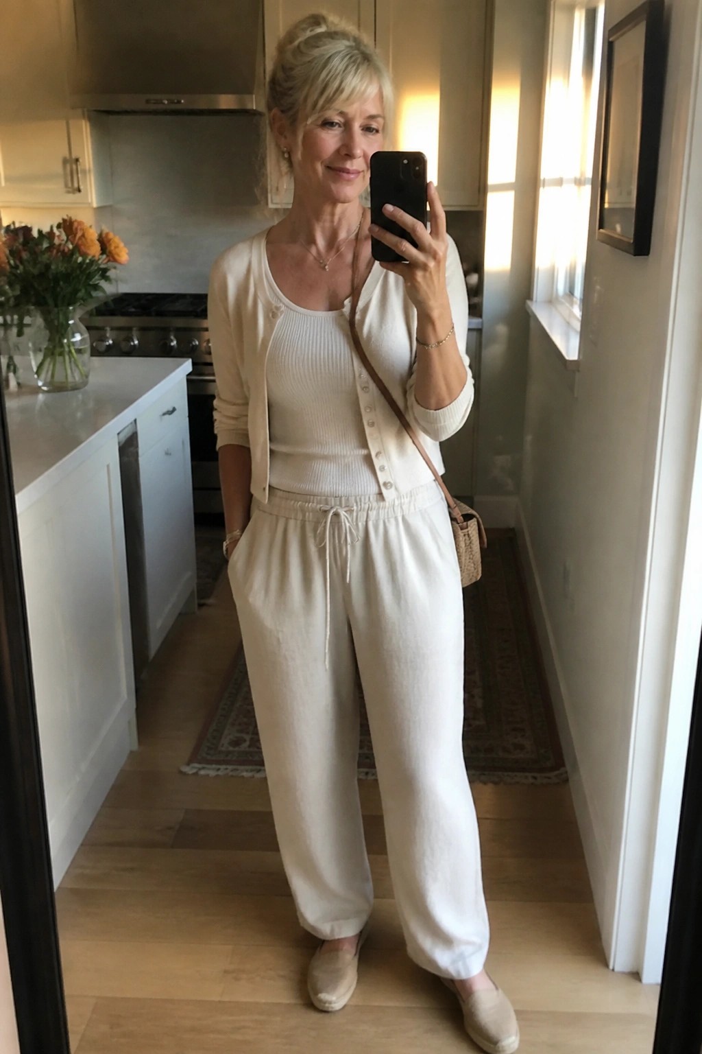 Woman in cream cardigan and wide pants taking mirror selfie in kitchen