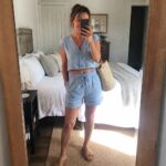 Woman over 50 wearing a light blue linen cropped button tank and matching drawstring shorts with tan sandals and a woven tote bag in a bedroom mirror selfie.