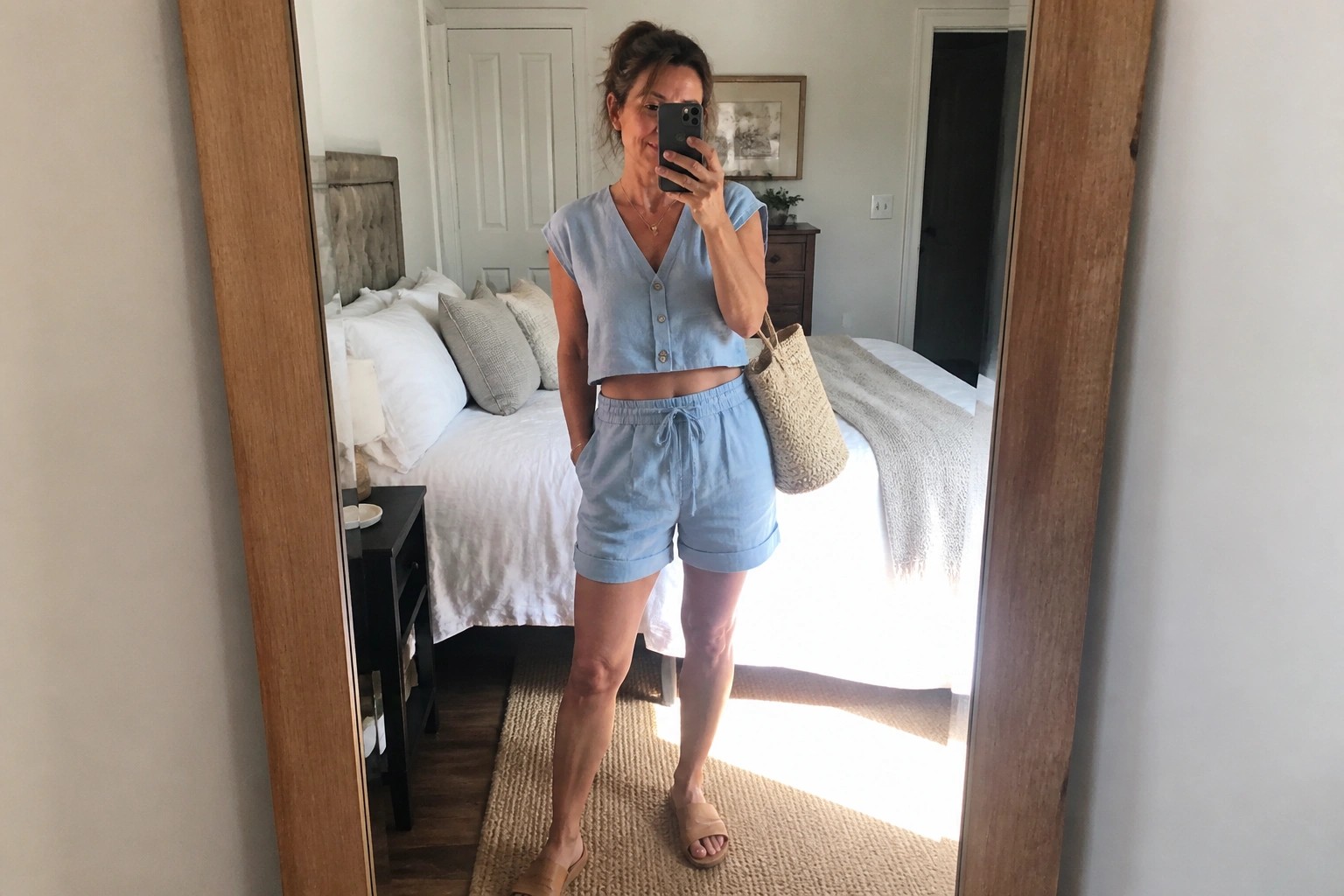 Woman over 50 wearing a light blue linen cropped button tank and matching drawstring shorts with tan sandals and a woven tote bag in a bedroom mirror selfie.