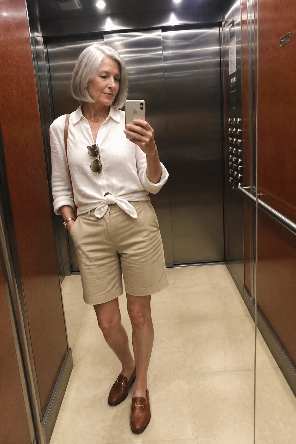 Woman with gray bob taking selfie in elevator wearing white shirt and beige shorts