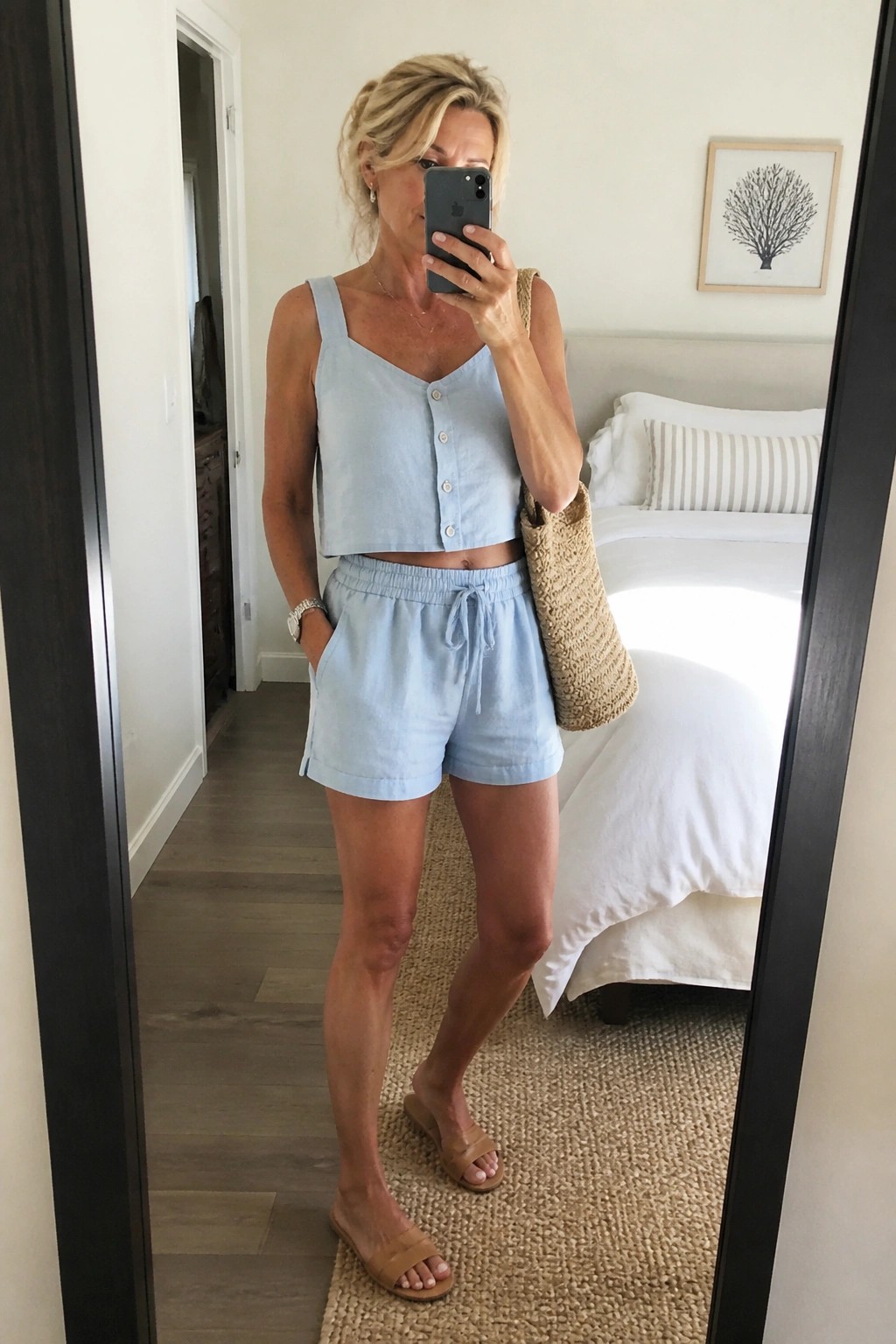Woman over 50 wearing a light blue linen cropped button tank and matching drawstring shorts with tan sandals and a woven tote bag in a bedroom mirror selfie.