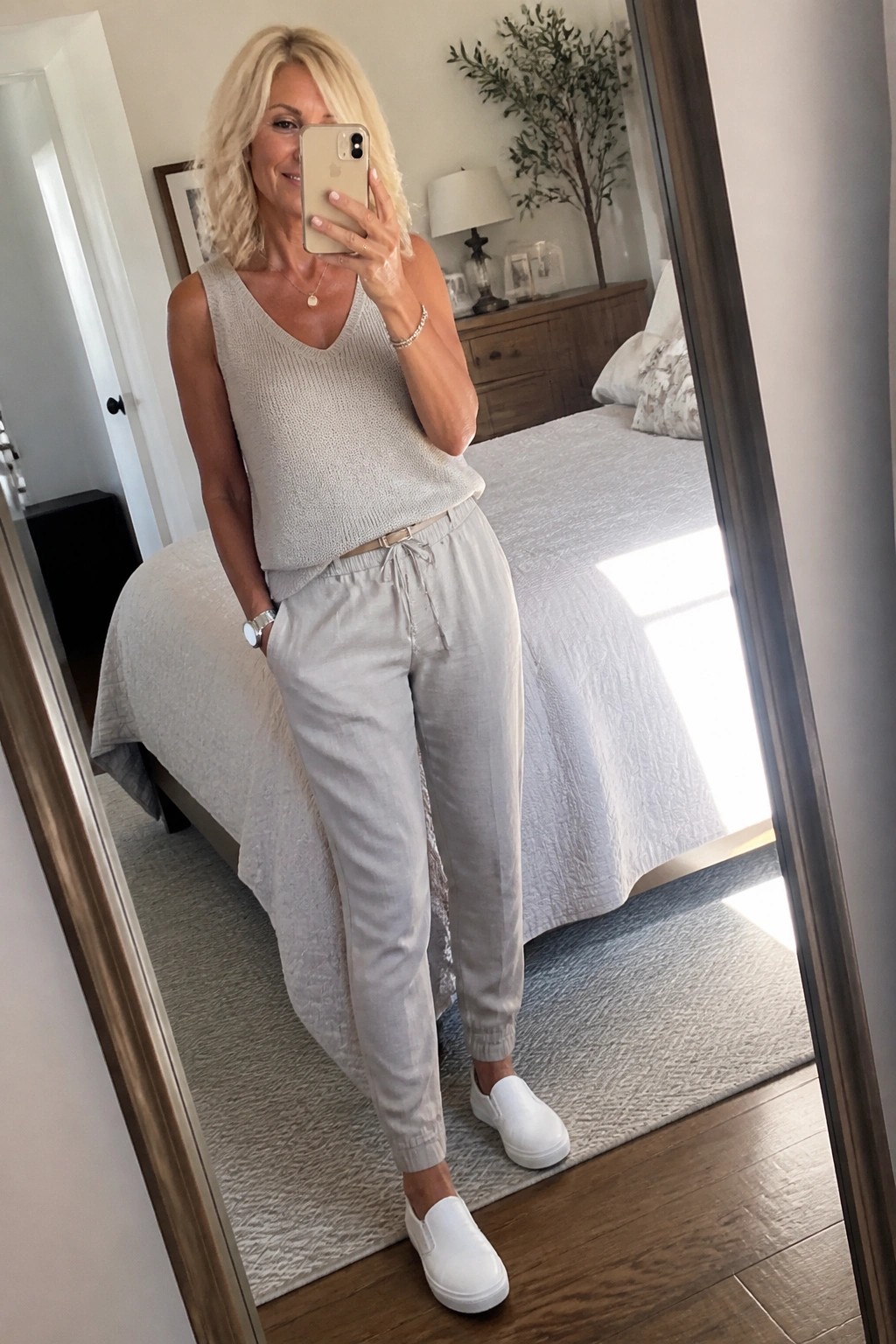 Blonde woman in beige knit top and joggers taking bedroom mirror selfie