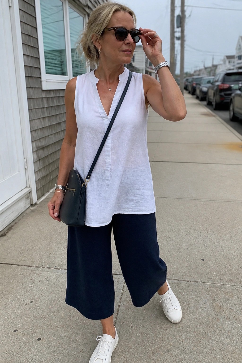 Blonde woman in white sleeveless top and navy culottes adjusts sunglasses on sidewalk.