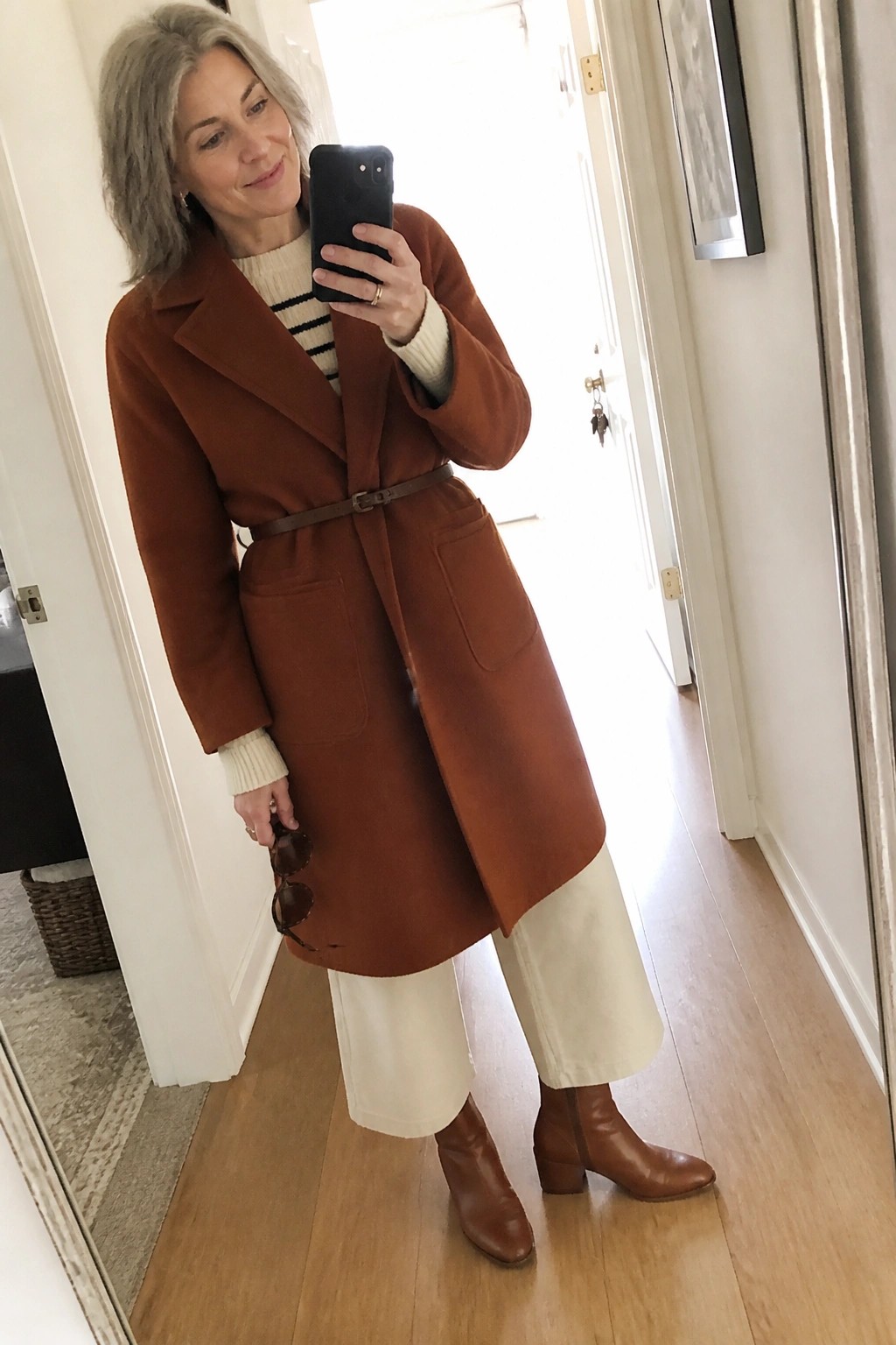 Gray-haired woman in rust coat and white pants taking mirror selfie