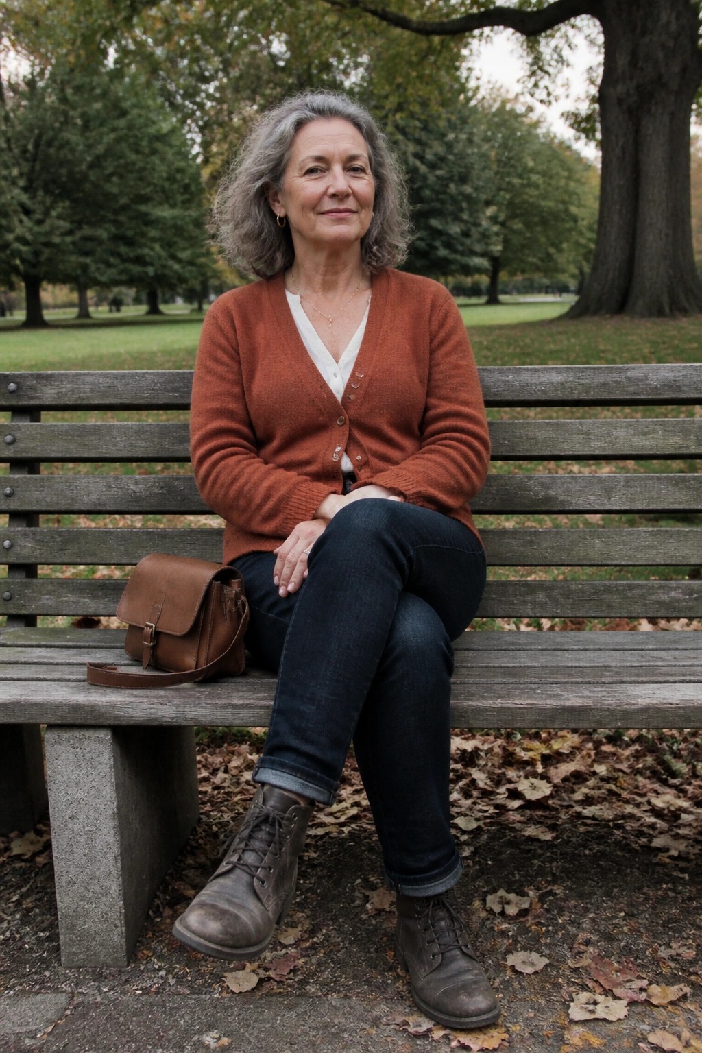Gray-haired woman in rust cardigan and jeans on leafy park bench with brown leather bag